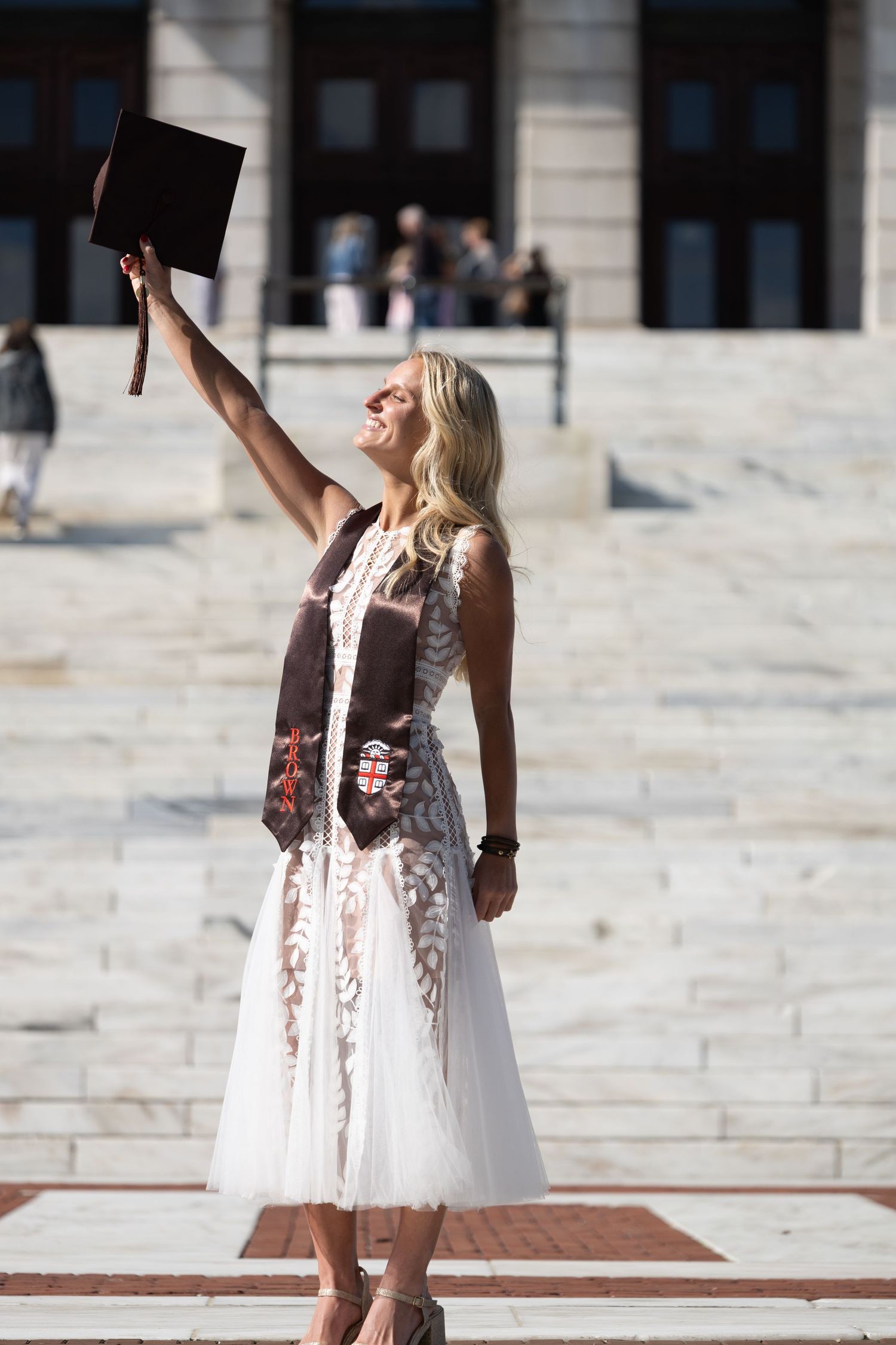 Blonde woman in white dress and graduation sash raises mortarboard cap triumphantly on university steps.