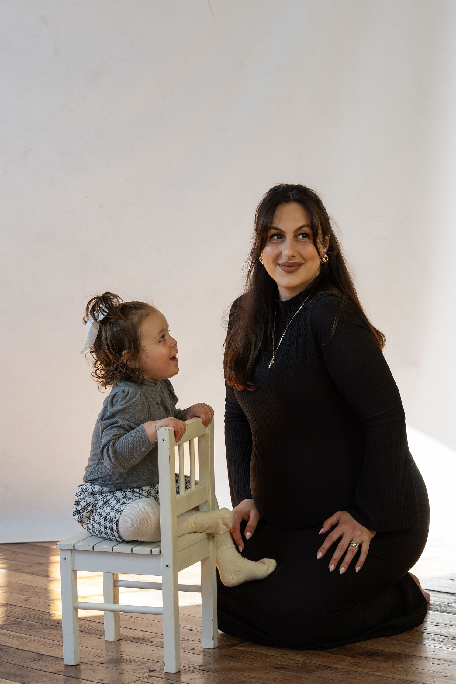 Pregnant woman in black dress sits on floor smiling with toddler girl on small white chair in studio.