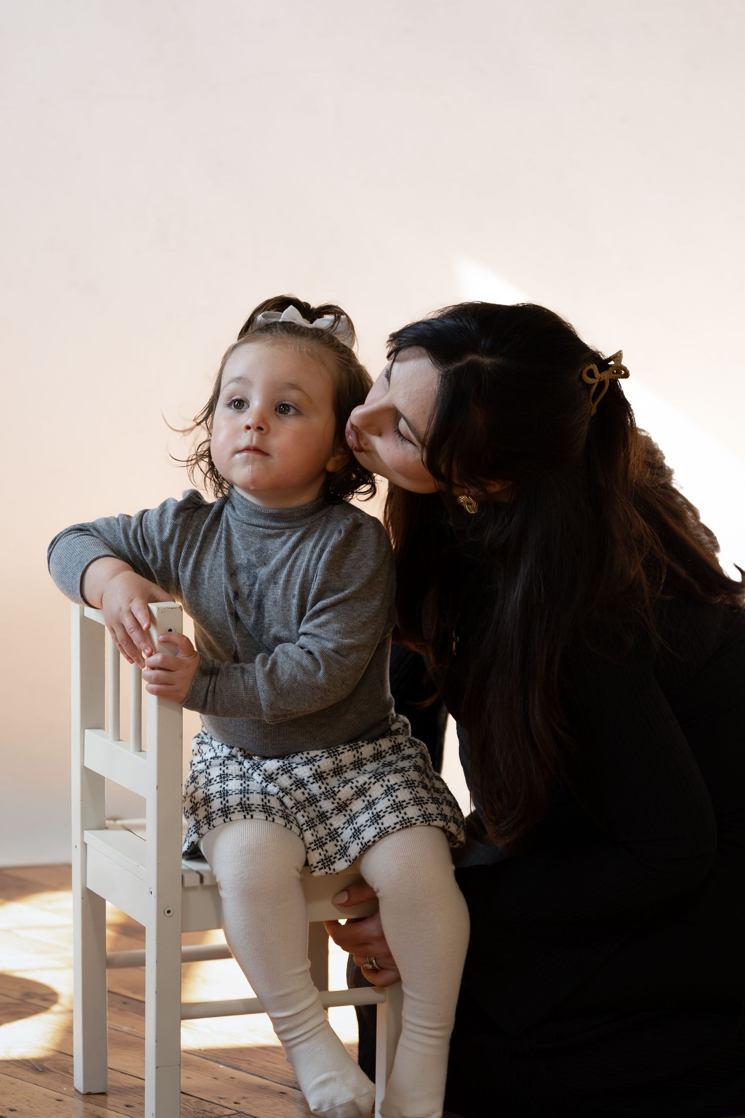 A dark-haired woman kisses a toddler girl in a gray top and plaid skirt sitting on a white chair.