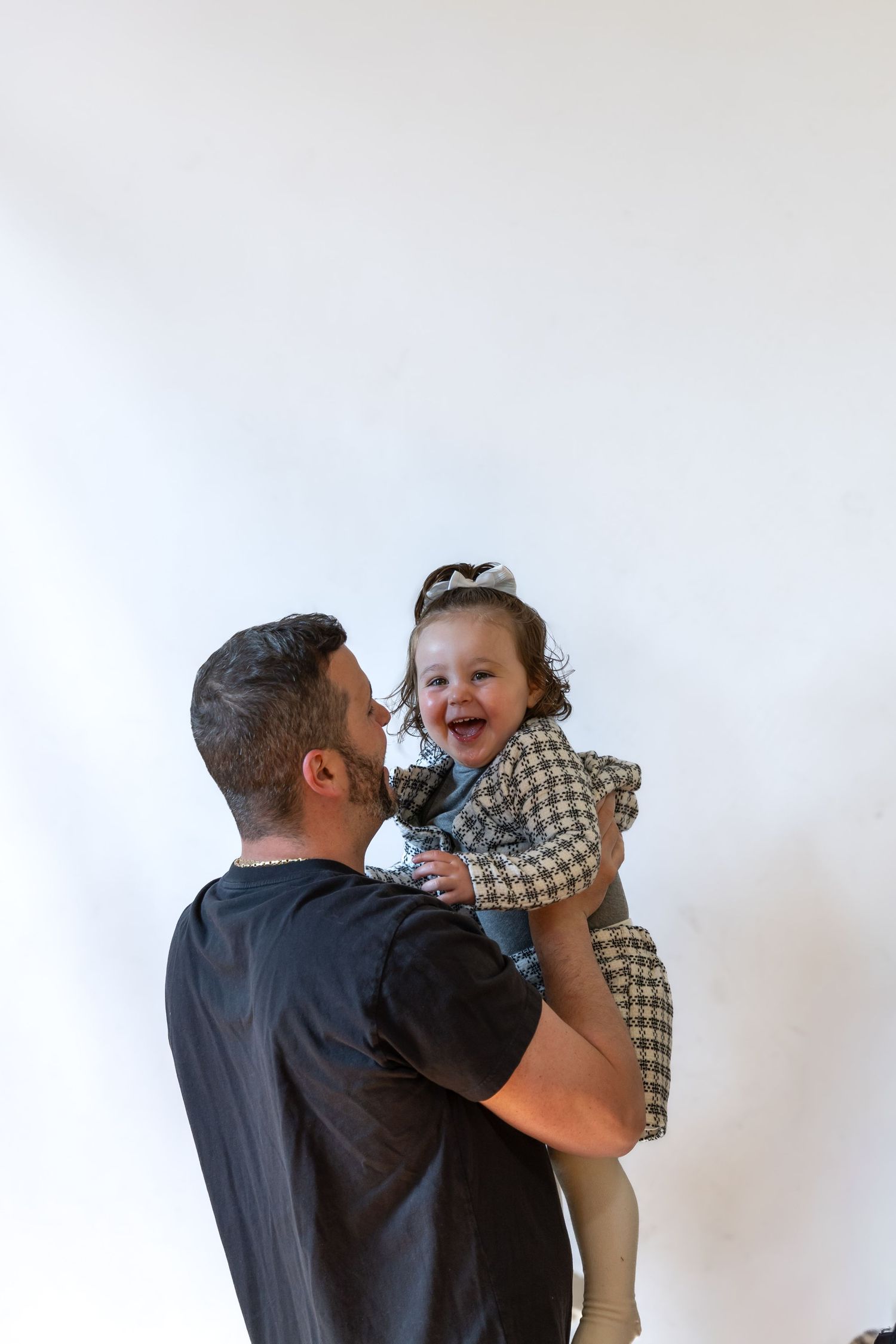 A joyful toddler girl in a plaid dress laughs while being lifted by a man in a black t-shirt against a white background.