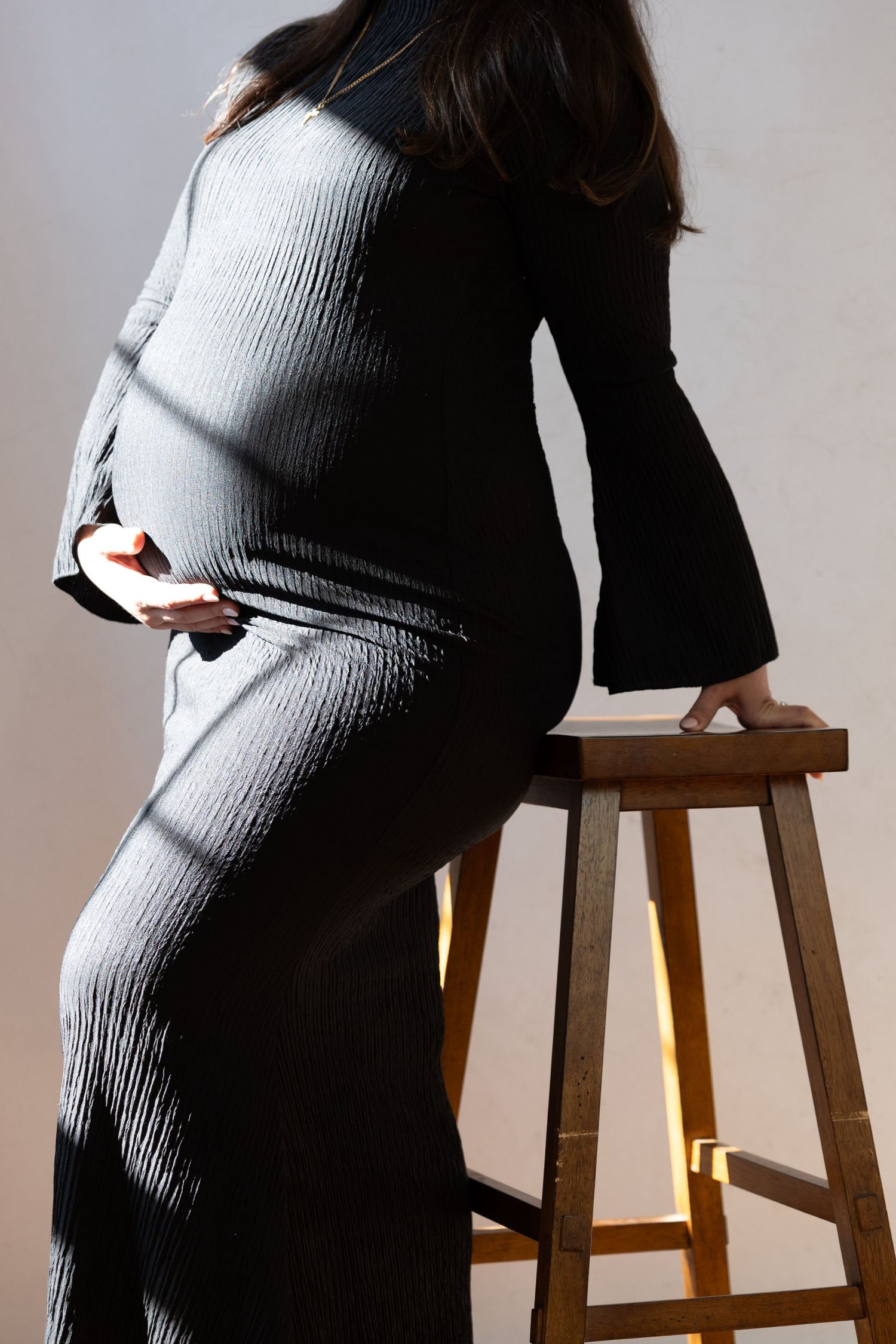 Pregnant woman in black ribbed dress holding baby bump, leaning on wooden stool in studio. Light from outdoors shining through window.