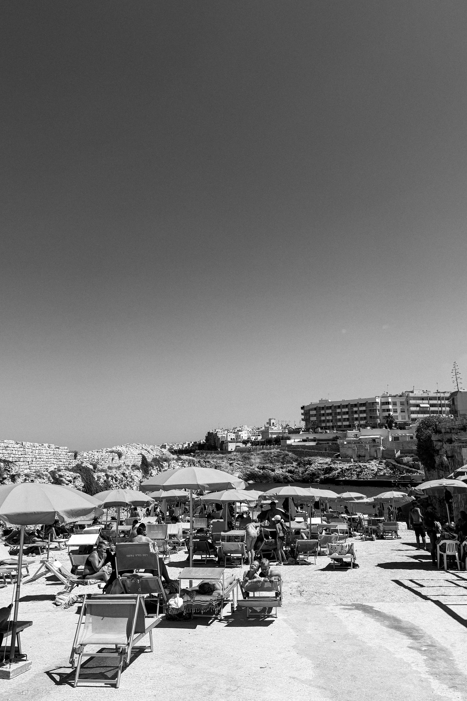 Beach umbrellas and lounge chairs line a sunny Mediterranean beach with buildings in the background.