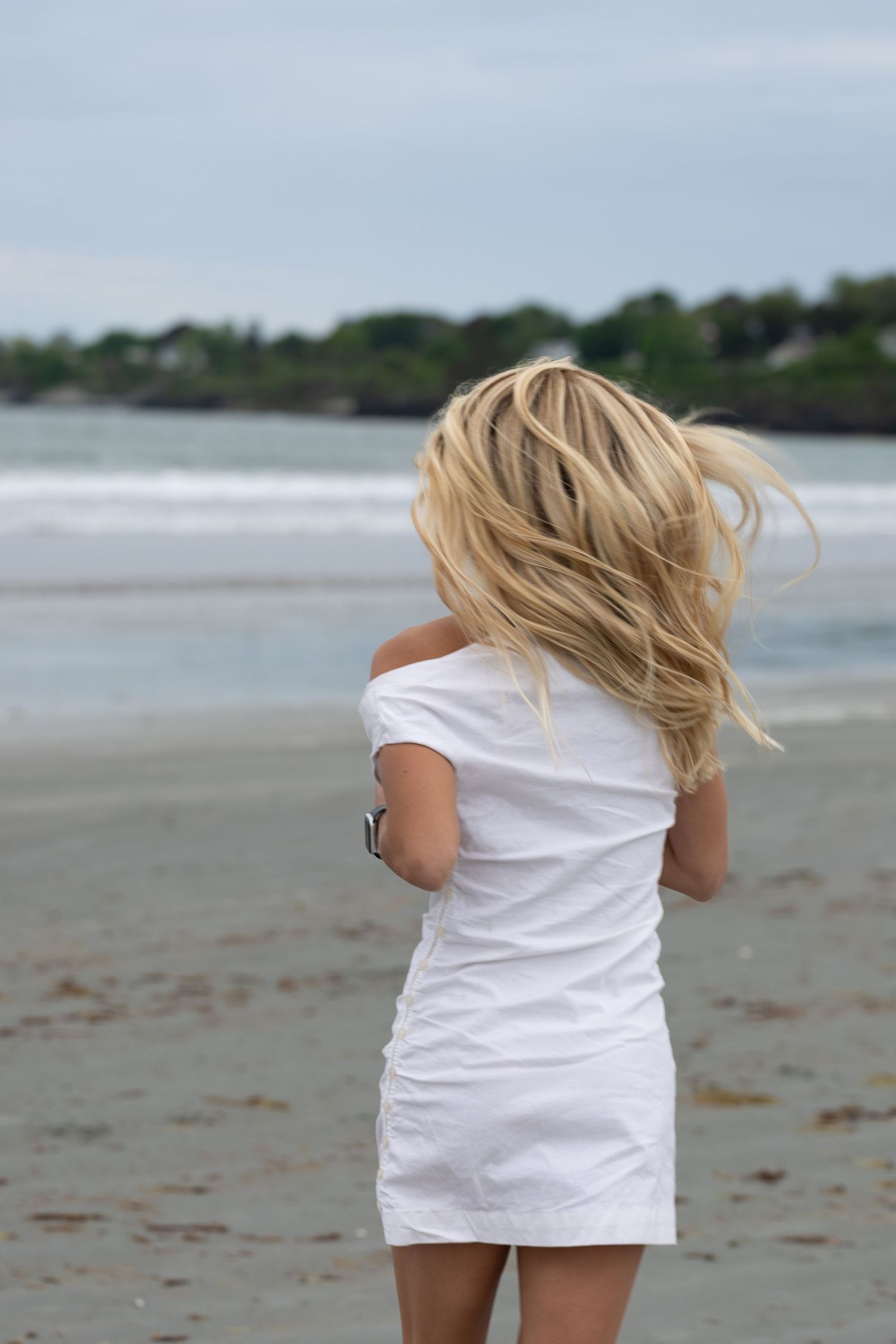 Blonde woman in white dress standing on a beach with hair blowing in the wind.