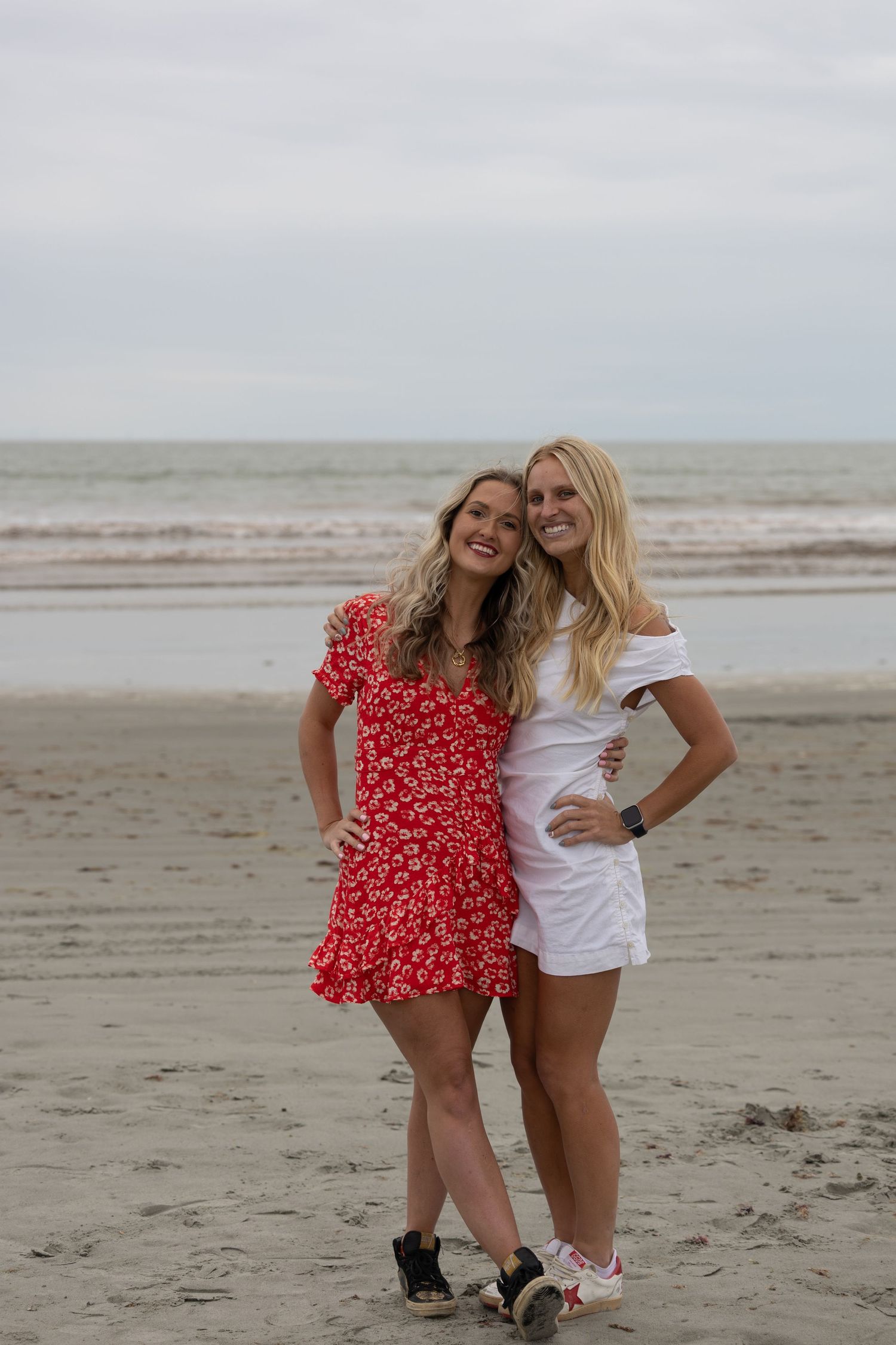 Two blonde women in red floral and white dresses posing together on a sandy beach with ocean waves behind them.