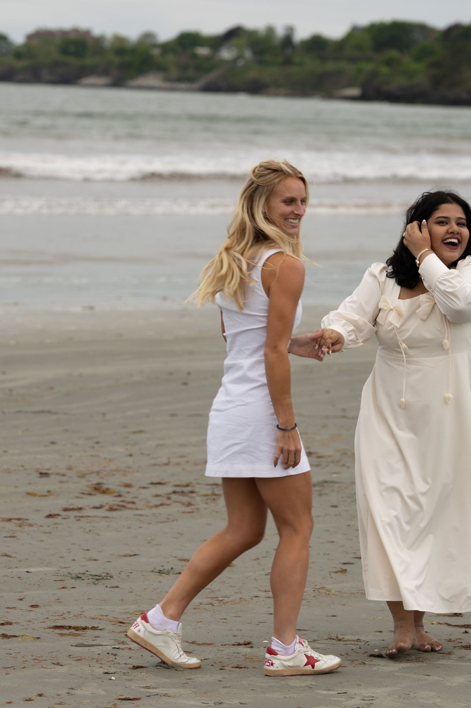 Two women in white dresses walk hand-in-hand on a sandy beach, laughing and enjoying a sunny day by the ocean.