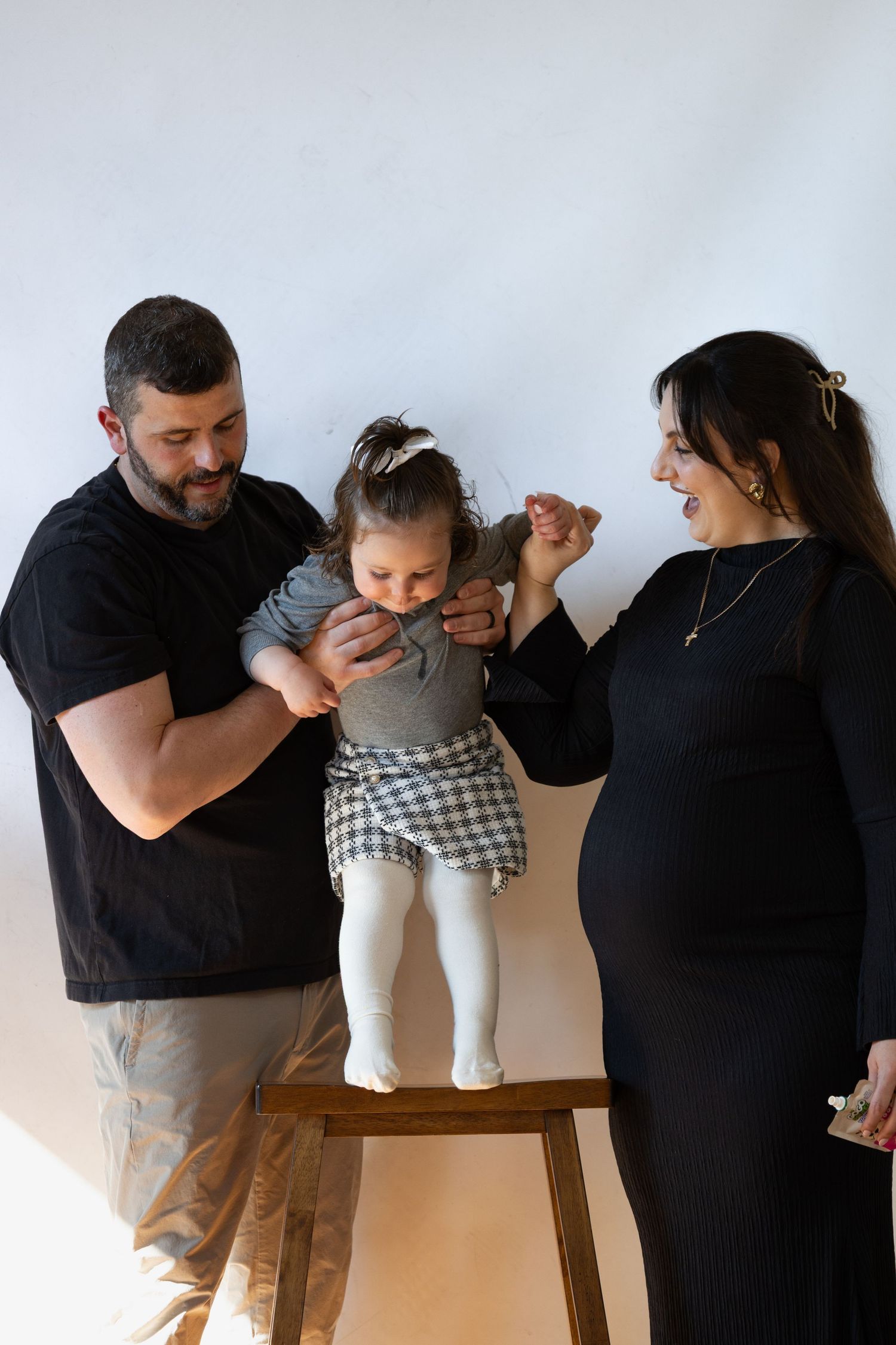 Parents in matching black outfits helping their young toddler daughter stand on a wooden stool against a white wall.