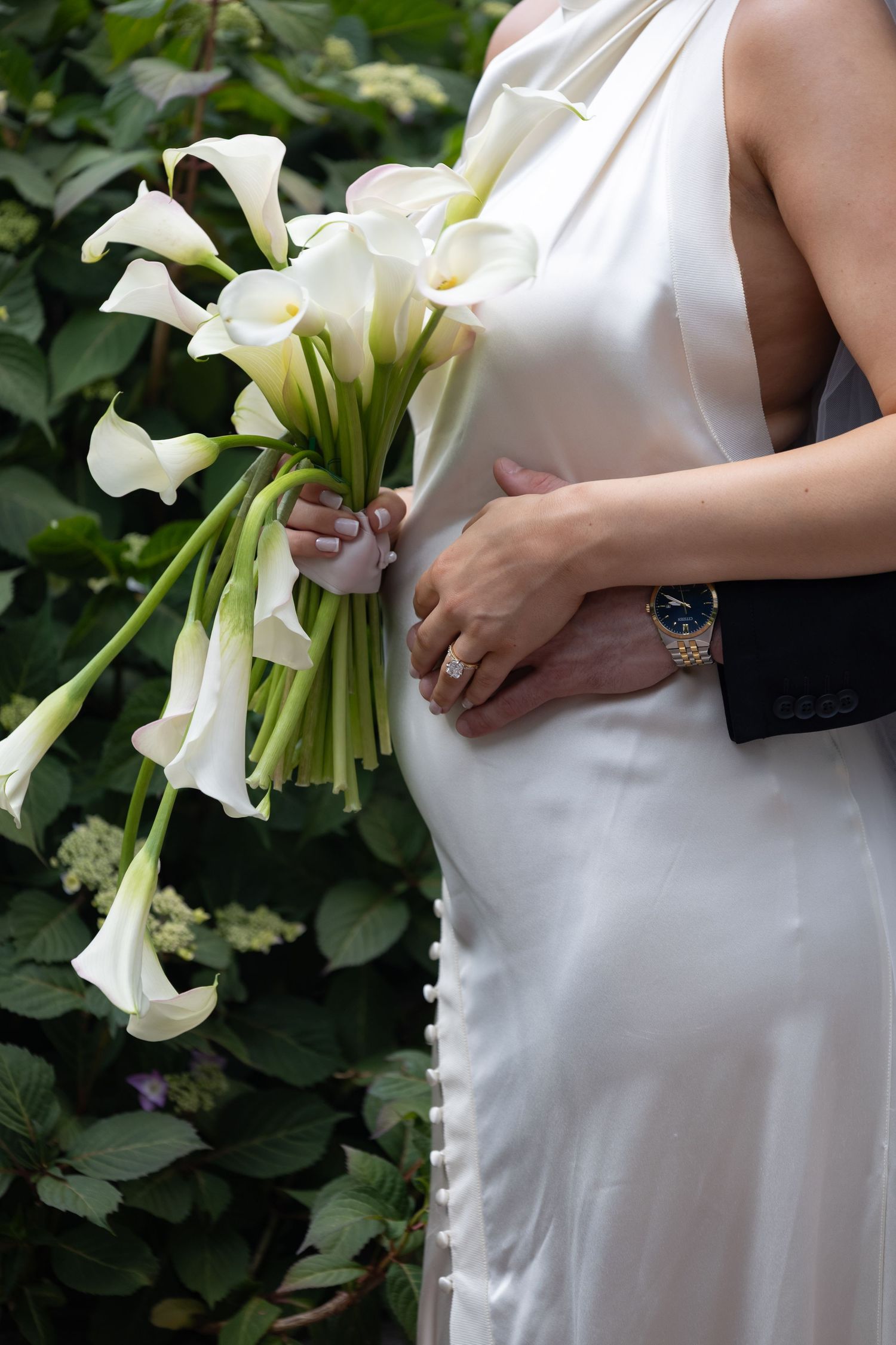 Pregnant bride in white dress holding calla lily bouquet with groom's hand on her belly outdoors.