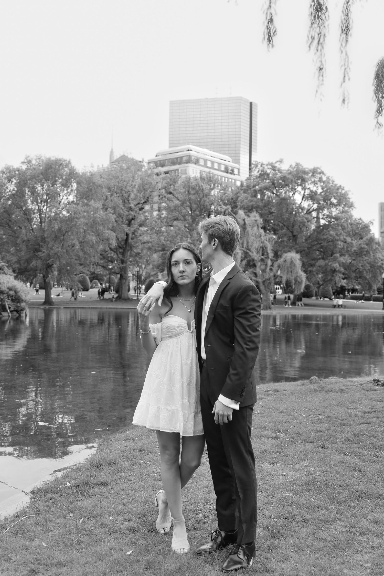 A couple poses romantically by a scenic lake with trees and buildings in the background.