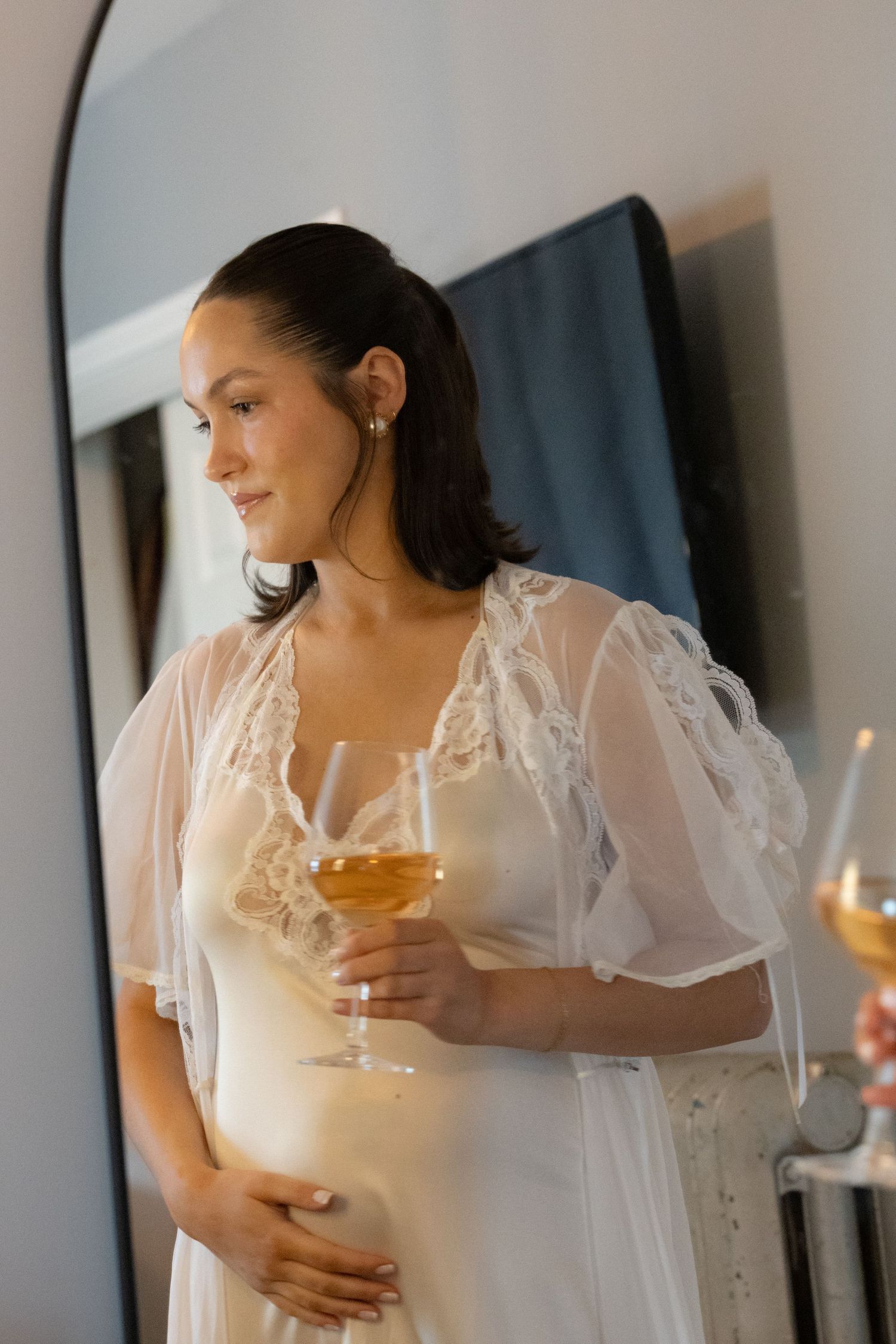 Pregnant woman in white lace maternity gown holding a wine glass, standing near a mirror in an elegant room.