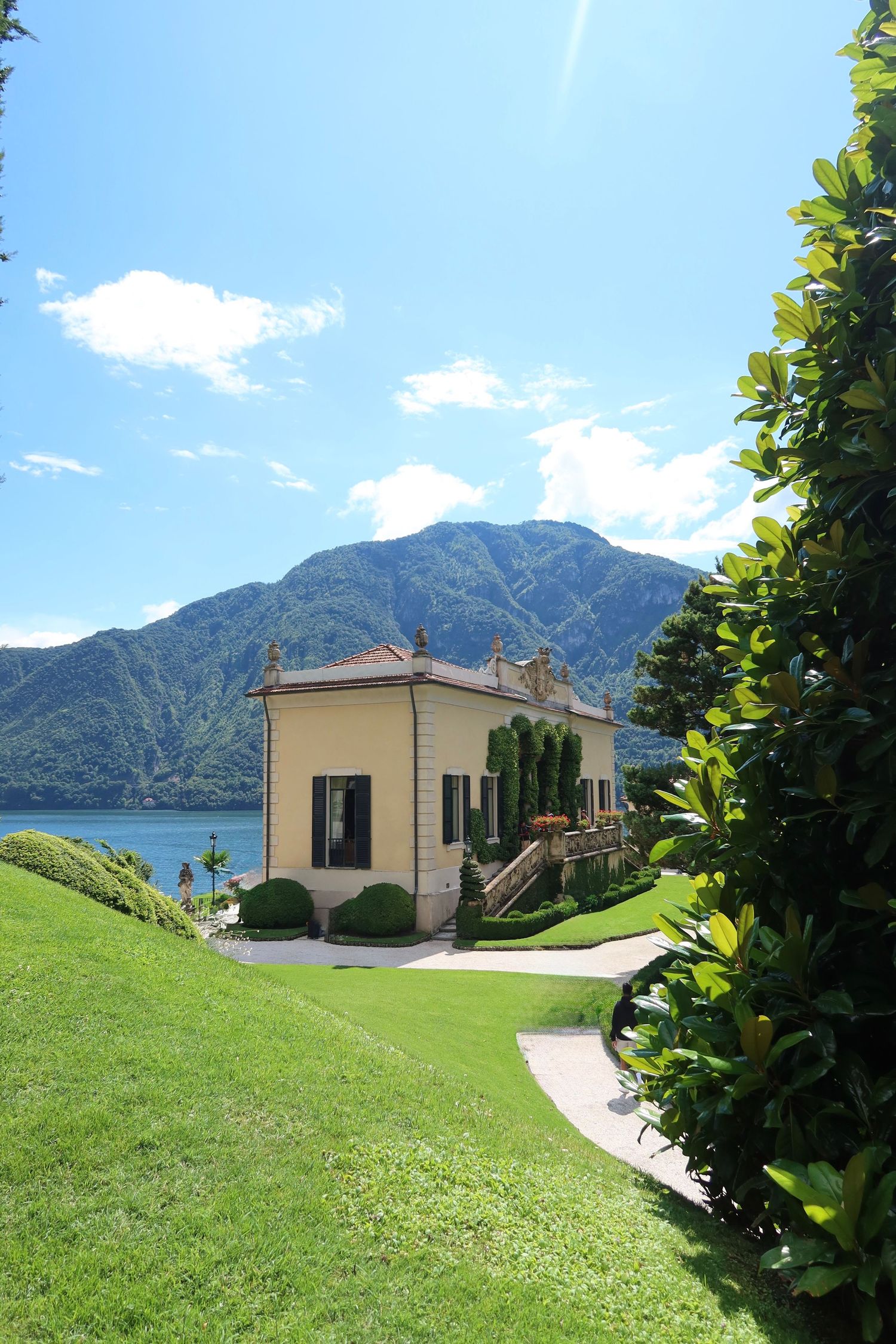 An elegant villa with cream-colored walls sits on a manicured lawn overlooking a mountain lake.