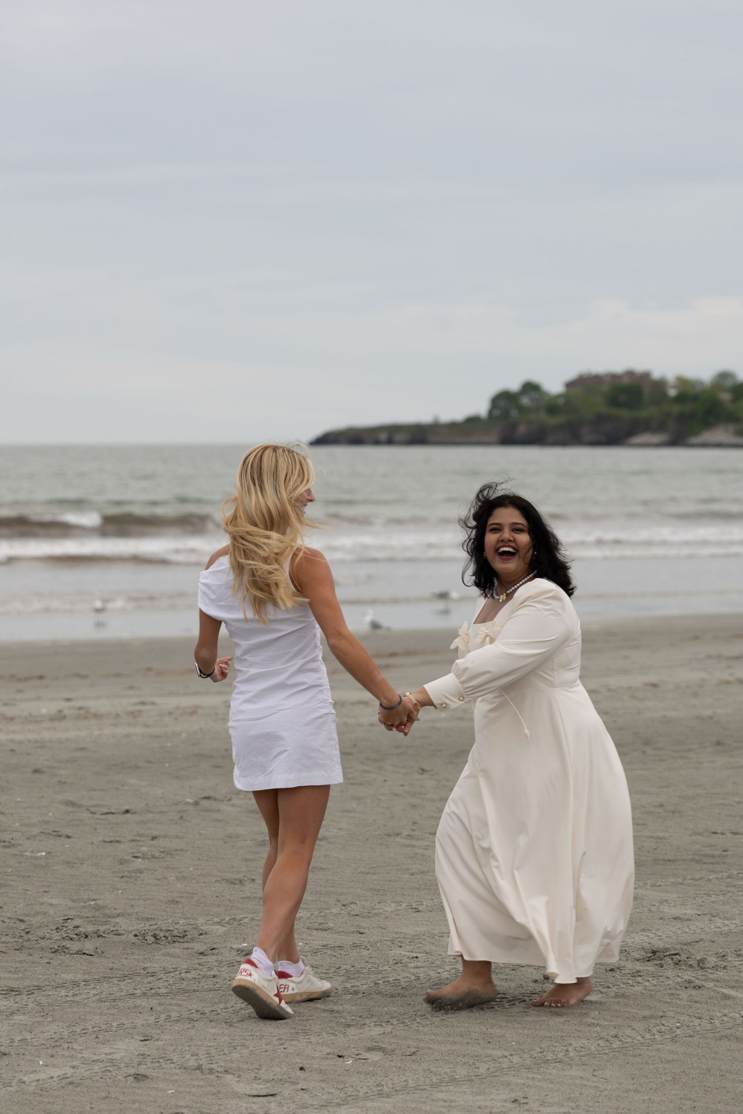 Two women in white dresses hold hands and laugh joyfully on a sandy beach with ocean waves in the background.