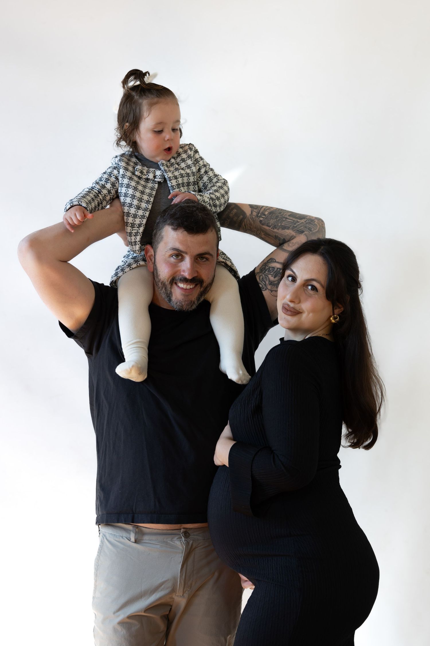 A smiling family with a tattooed dad carrying a toddler on his shoulders beside a pregnant mom in black.
