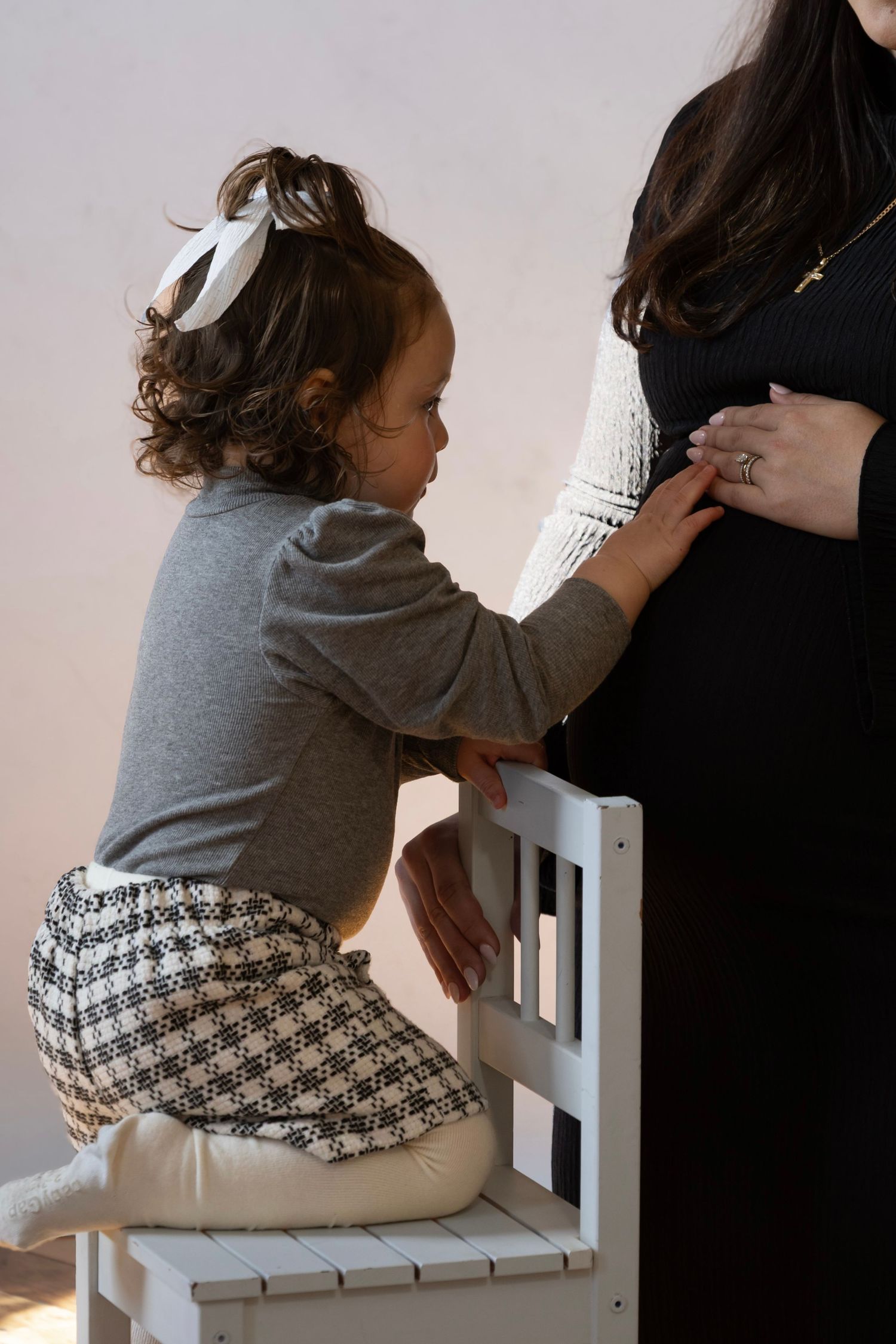 Young toddler girl kneeling on chair touching pregnant mother's belly in soft natural light.