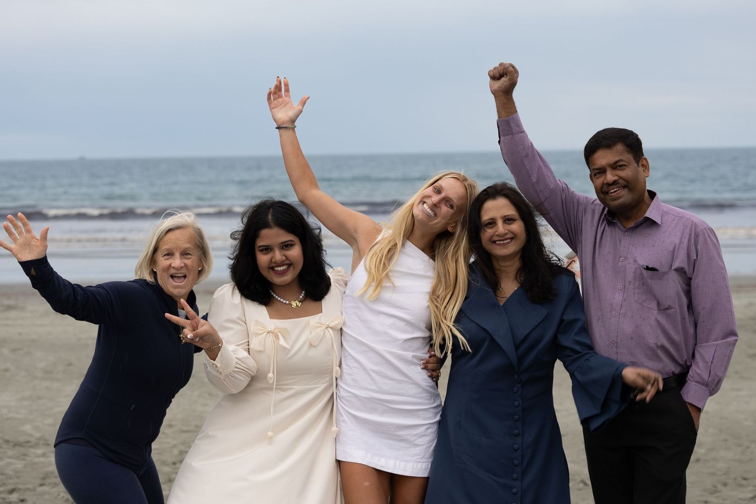 Five people celebrating on a beach, smiling and raising their arms in joy on a cloudy day.