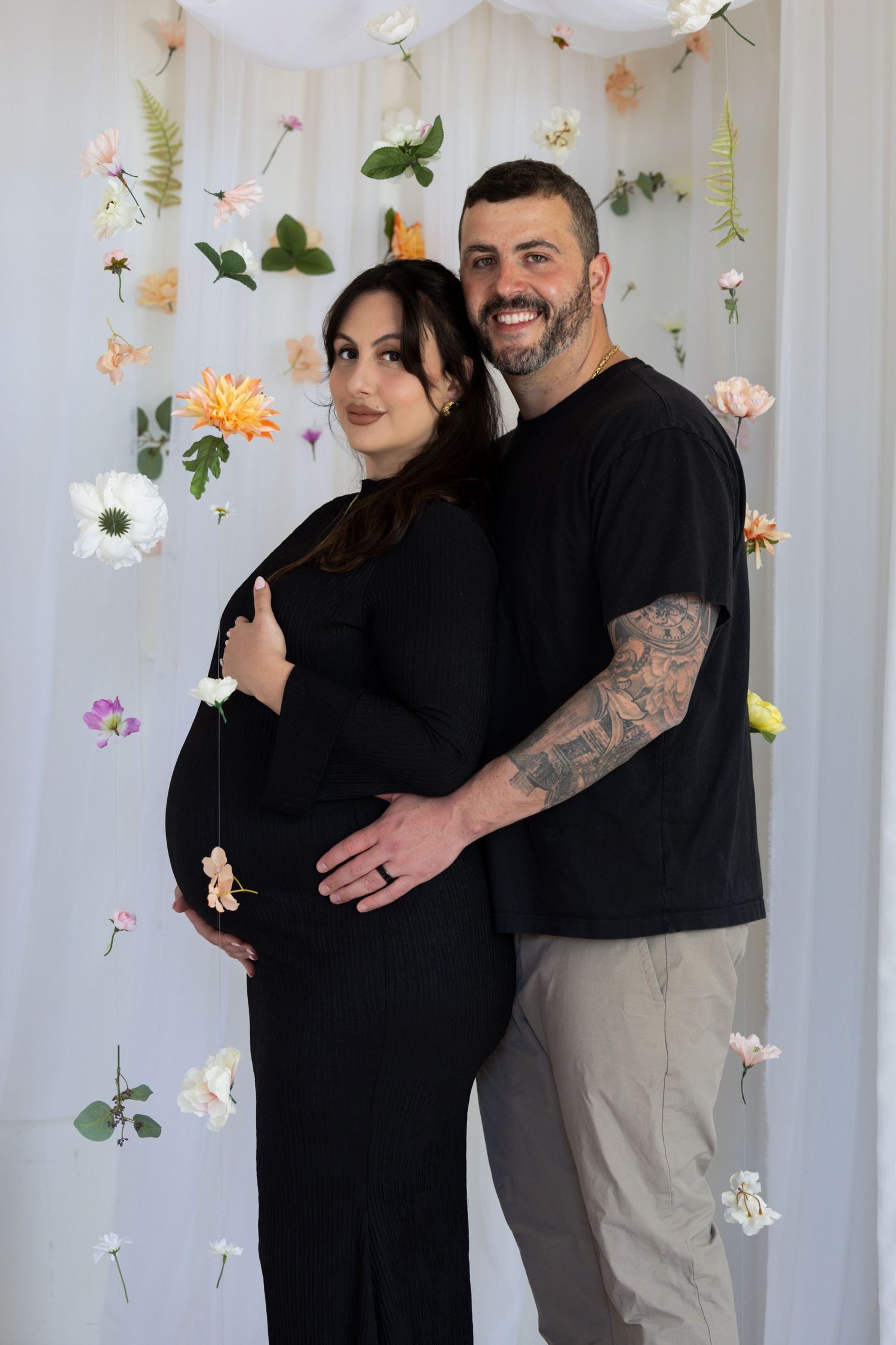 Smiling couple in black outfits pose for maternity photo in front of floral backdrop, man embracing pregnant woman.