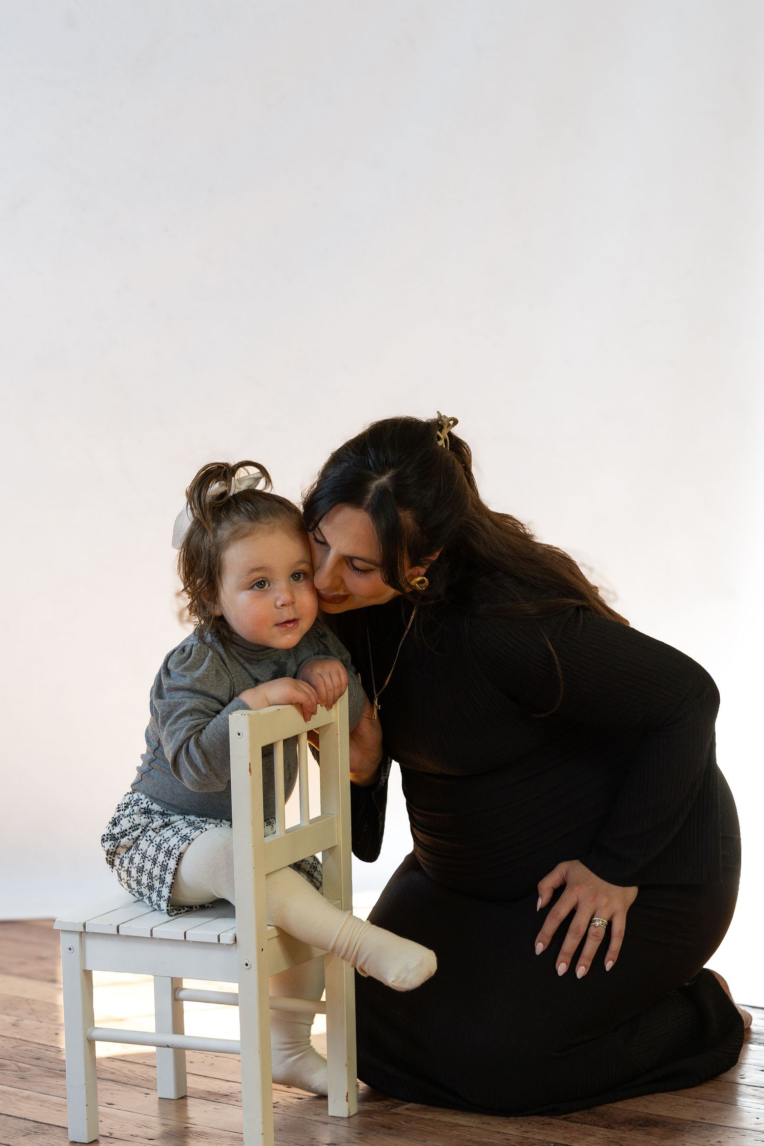 A woman in black dress kneeling beside a toddler sitting on a small white chair in a bright studio.