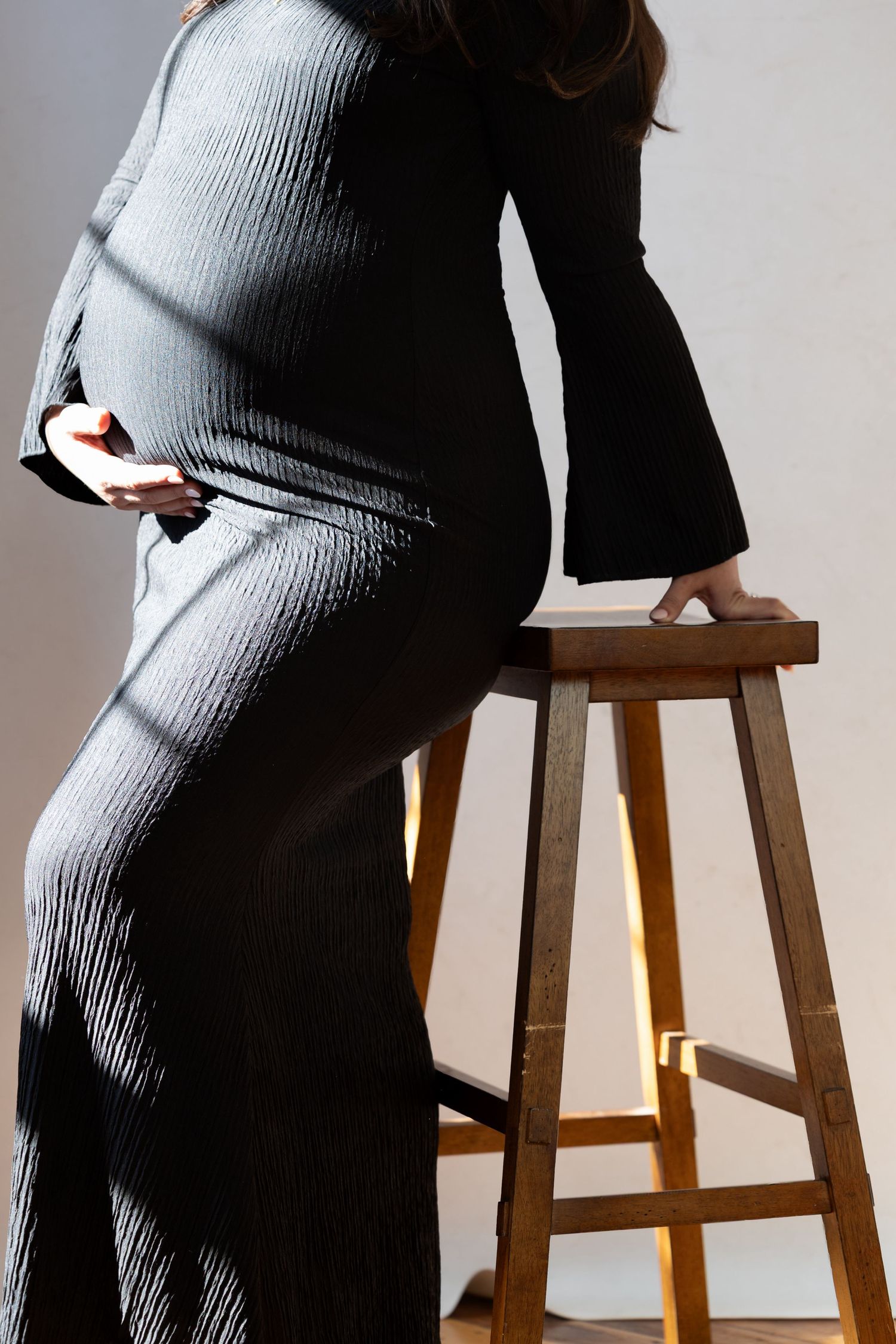 Side profile of pregnant woman in grey ribbed dress cradling bump beside wooden stool.