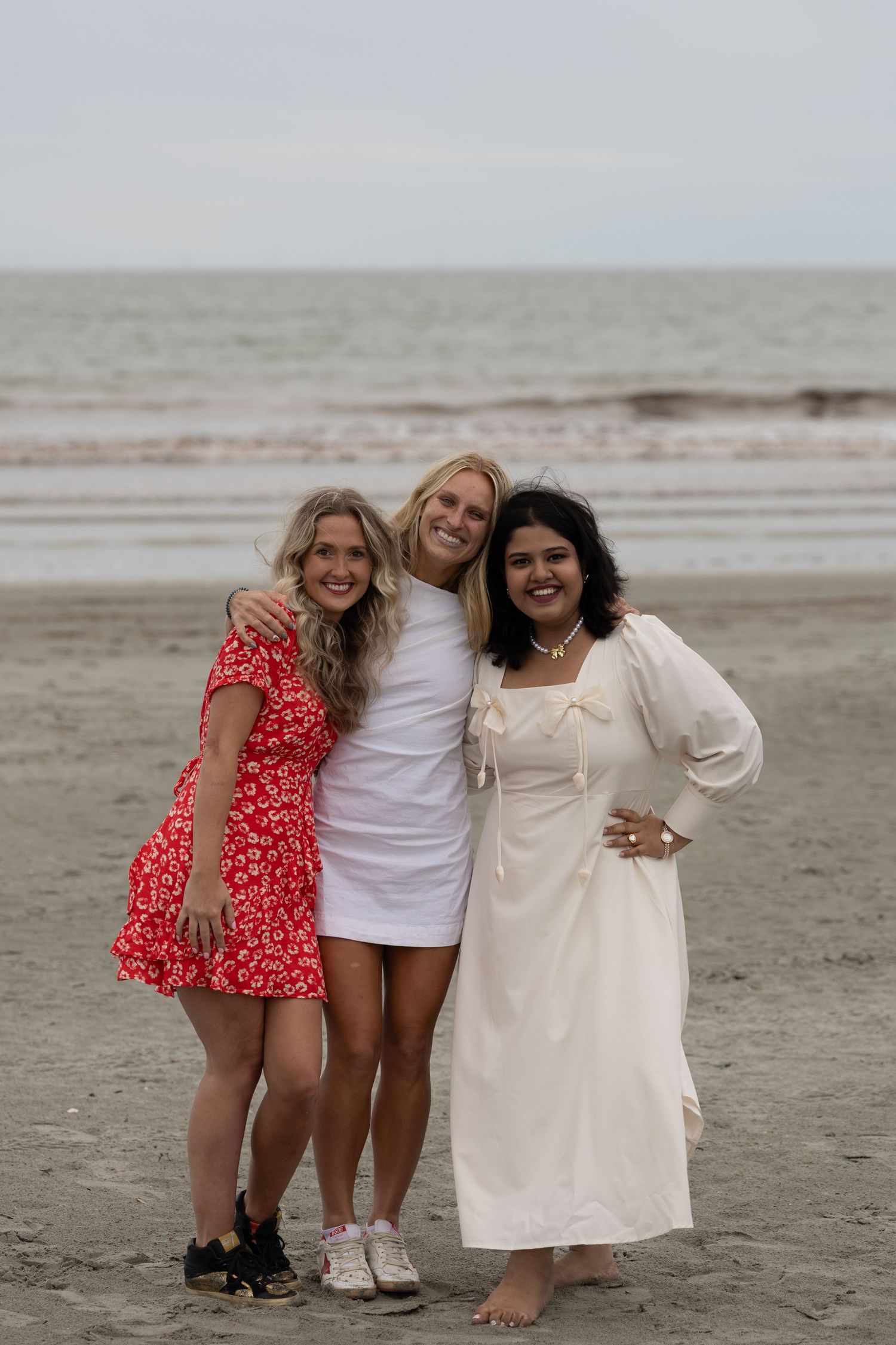 Three young women smiling and posing together on a sandy beach, wearing red floral, white mini, and cream maxi dresses.