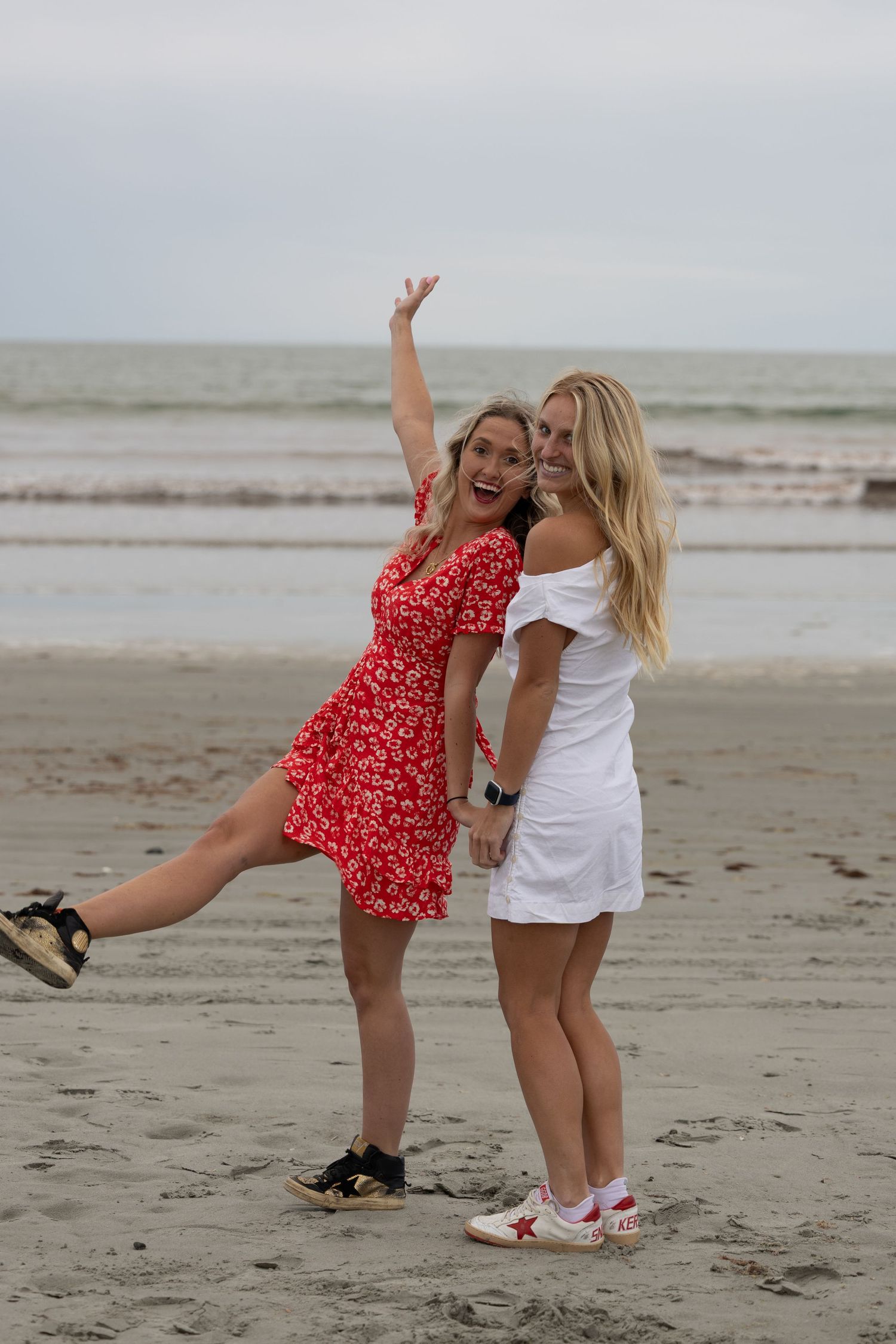 Two blonde women in red and white outfits posing playfully on a sandy beach with ocean waves behind them.