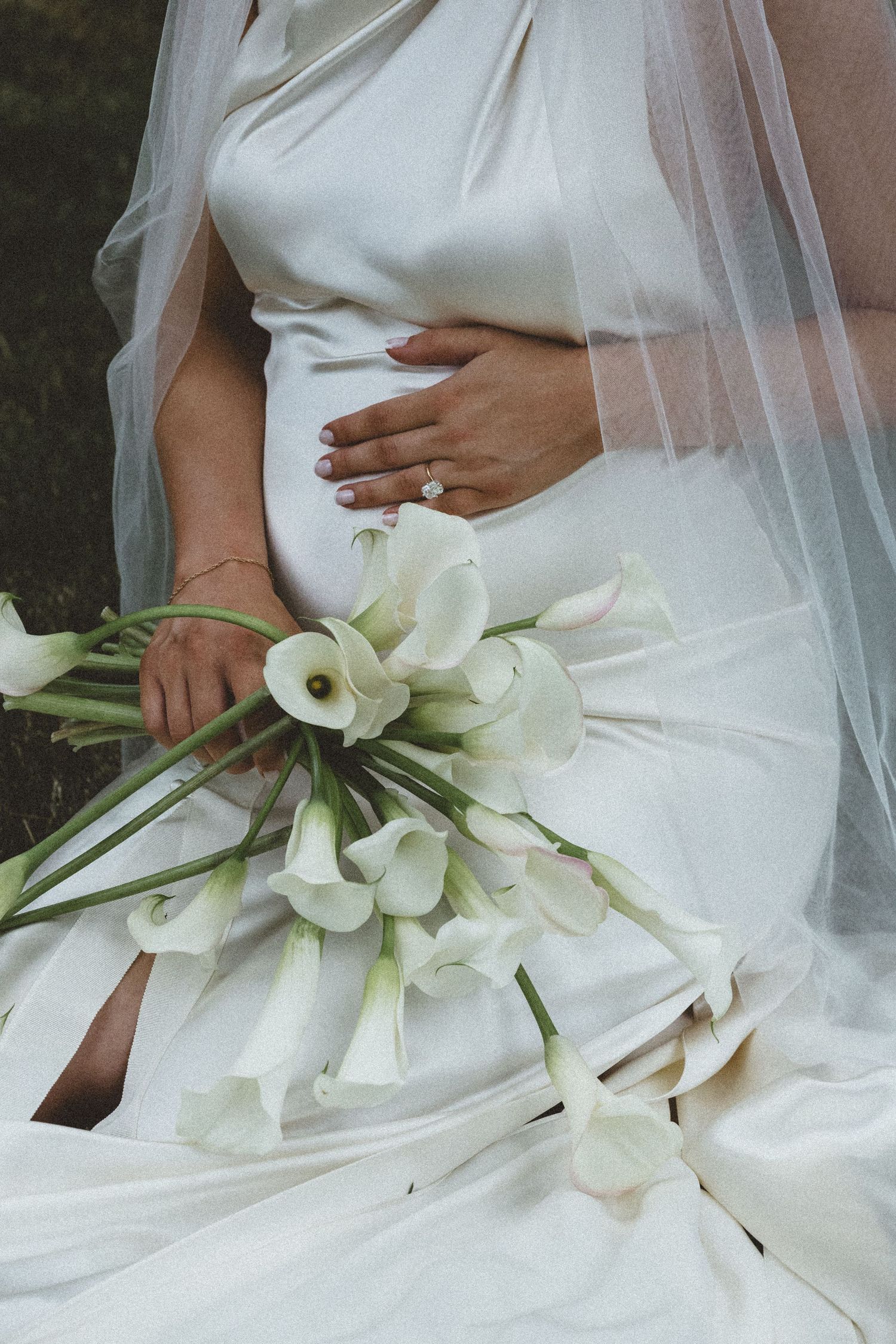 Pregnant bride in white silk wedding dress holding calla lily bouquet with diamond ring visible.