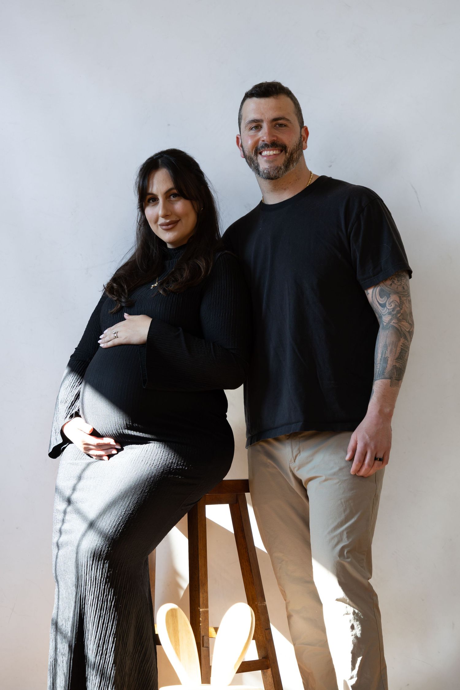 Smiling couple posing for maternity photo, woman in black outfit cradling baby bump, man with tattoo in black tee.