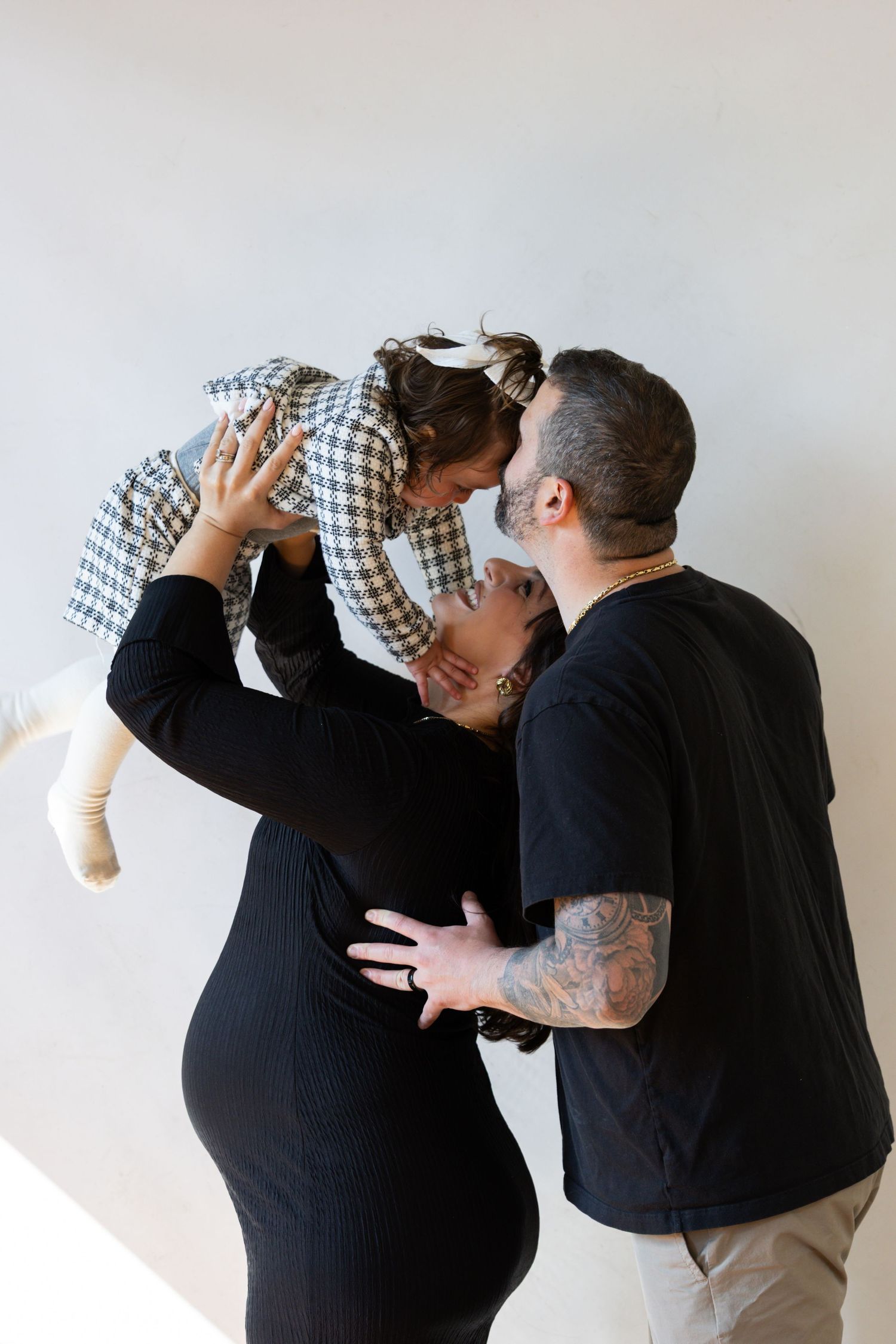 Expecting couple in black outfits lifting toddler daughter between them against white wall.