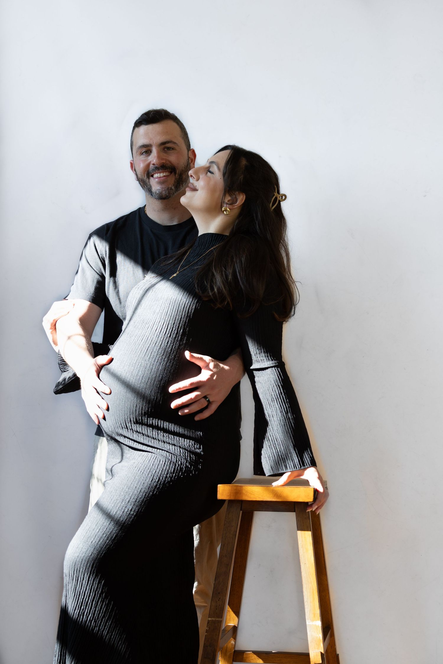 Happy couple embracing, pregnant woman in grey dress leaning on wooden ladder against white wall.