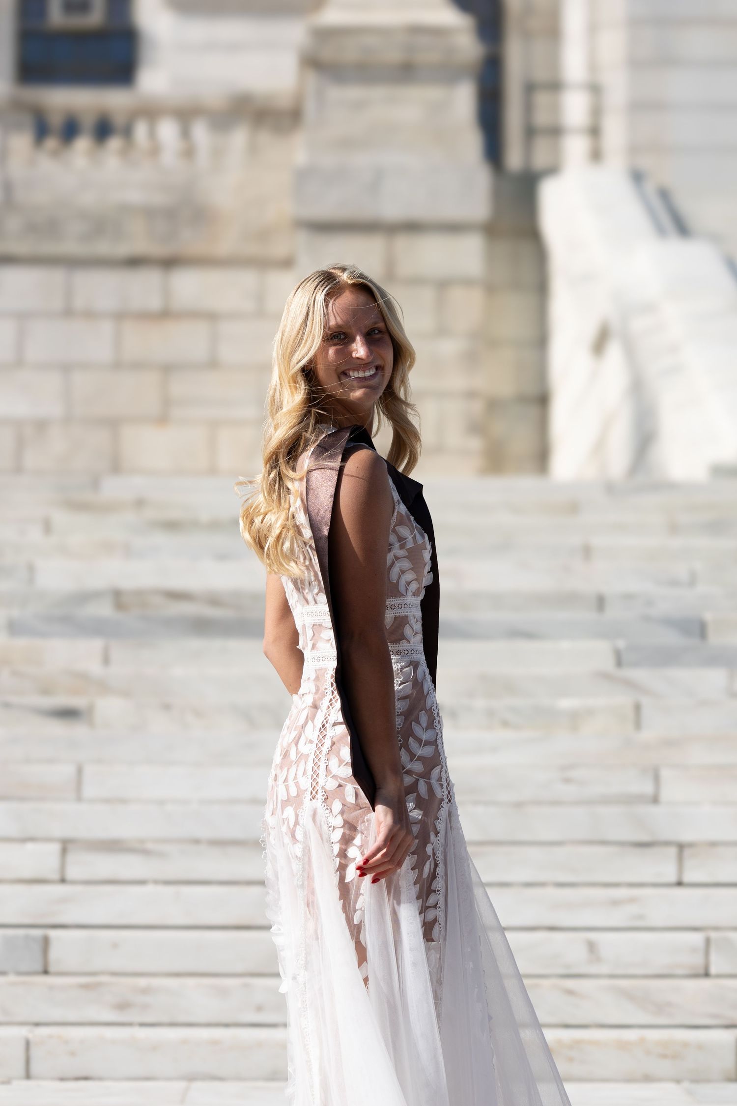 Blonde woman in elegant white lace backless gown posing on marble steps at a classical architectural landmark.