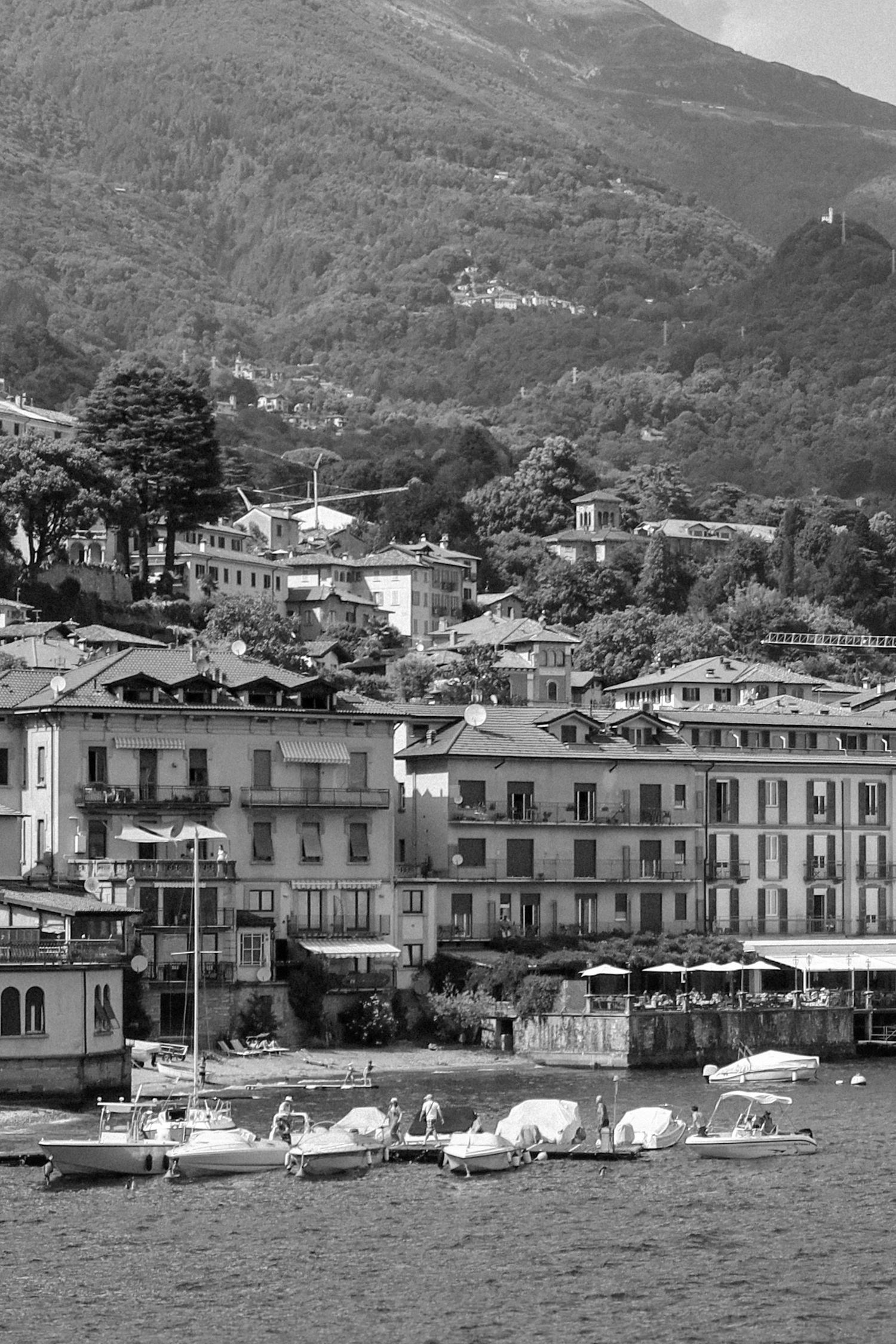 Lakeside village with white buildings and small boats moored along the waterfront against mountain backdrop.