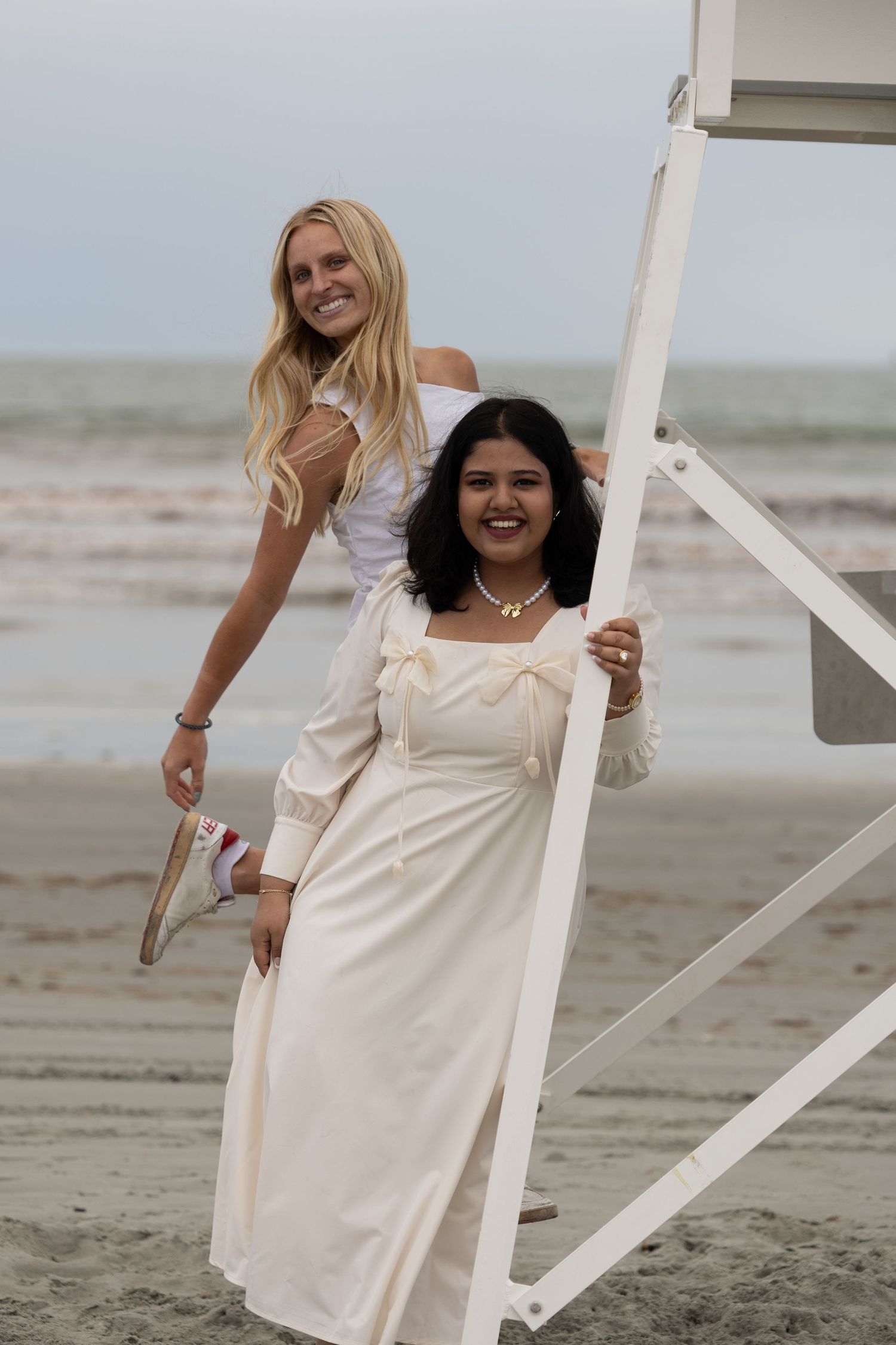 Two women in elegant dresses smiling by a lifeguard stand on a beach.