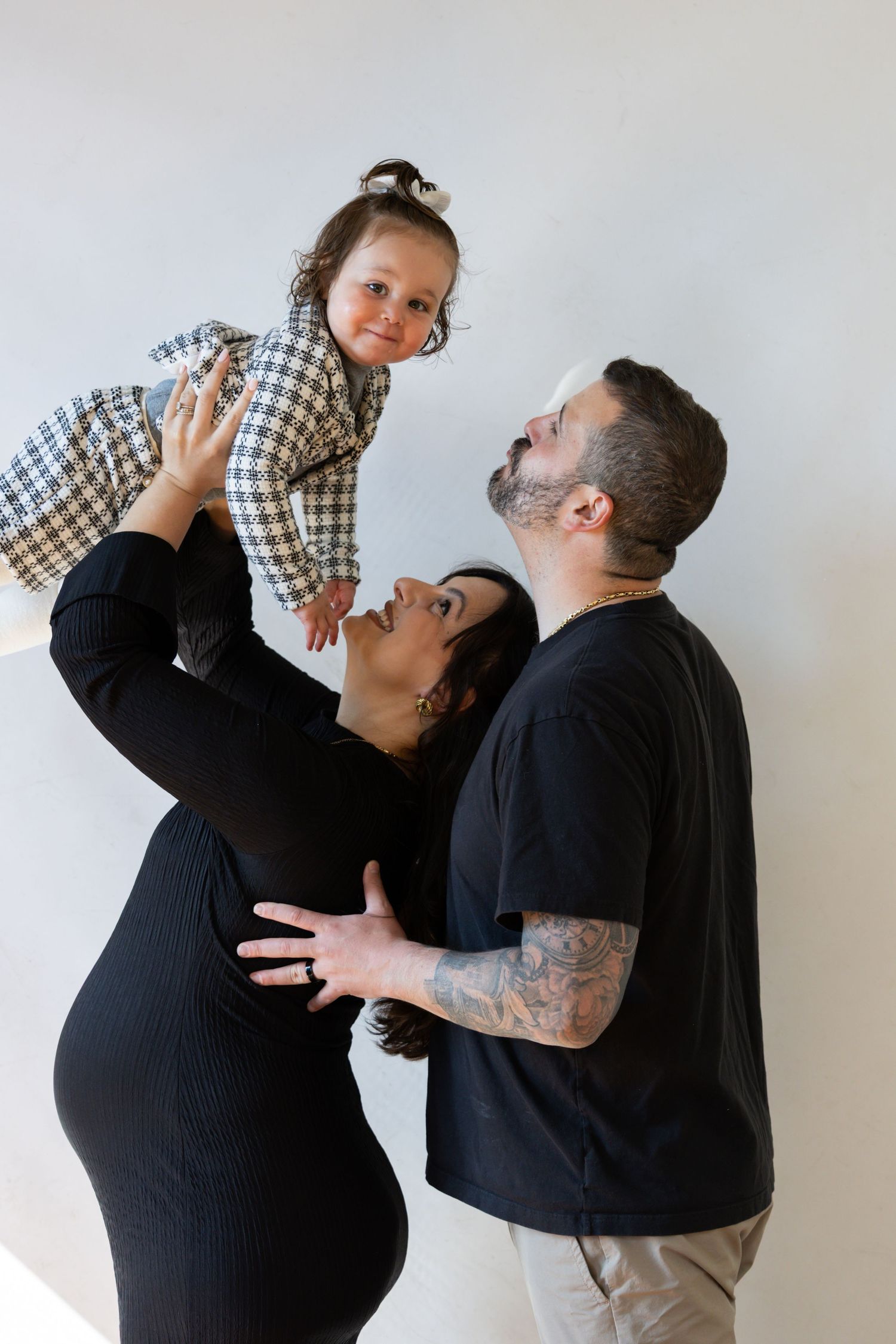 Pregnant mother lifting smiling toddler while father embraces her from behind during a maternity photo session.