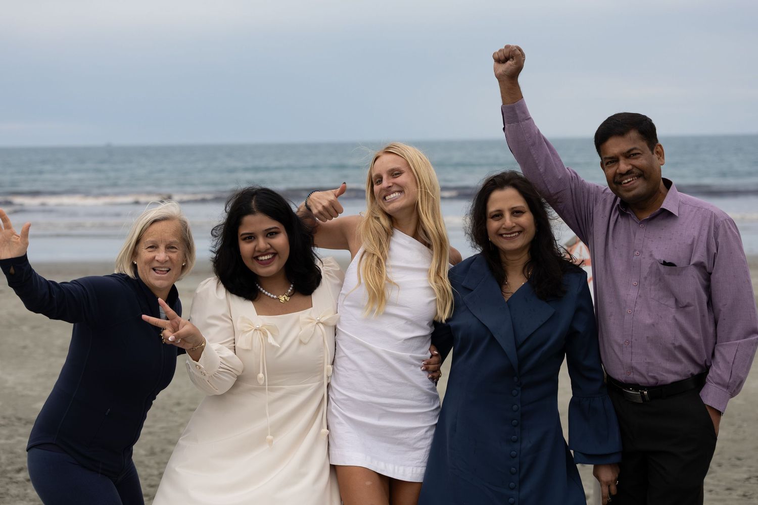 Five people posing joyfully on a beach, celebrating with raised arms and peace signs on a cloudy day.