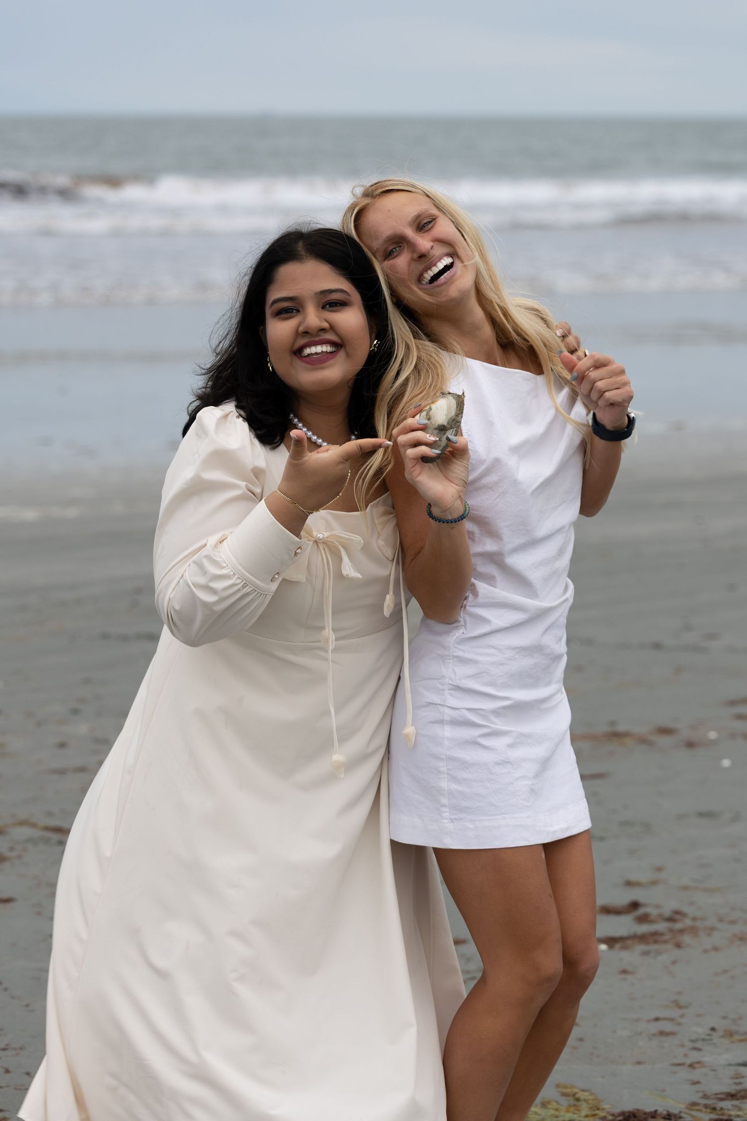 Two laughing women in white dresses pose playfully on a beach with ocean waves behind them.