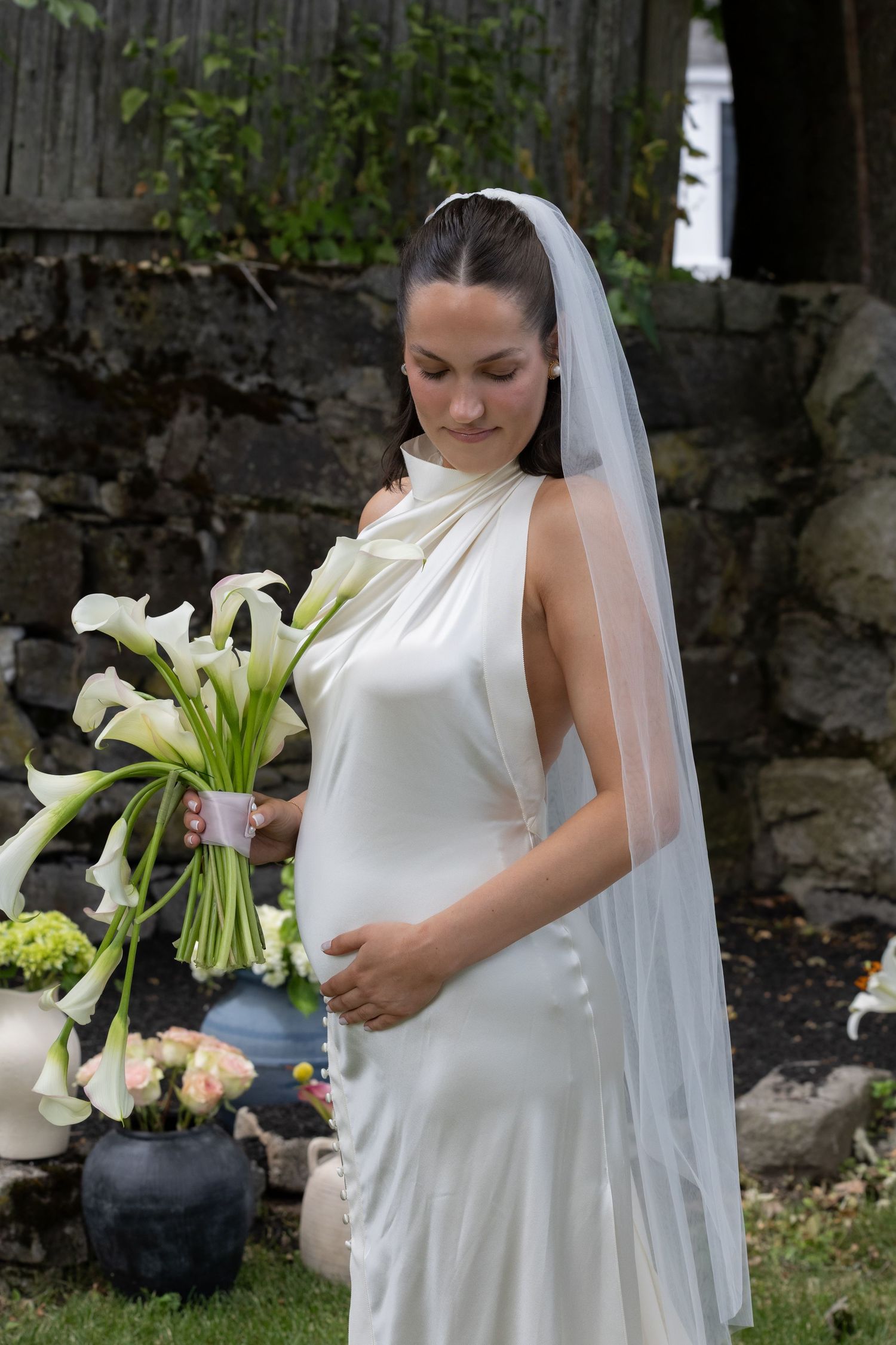 Pregnant bride in elegant white halter gown and veil gazing at calla lily bouquet in garden setting.