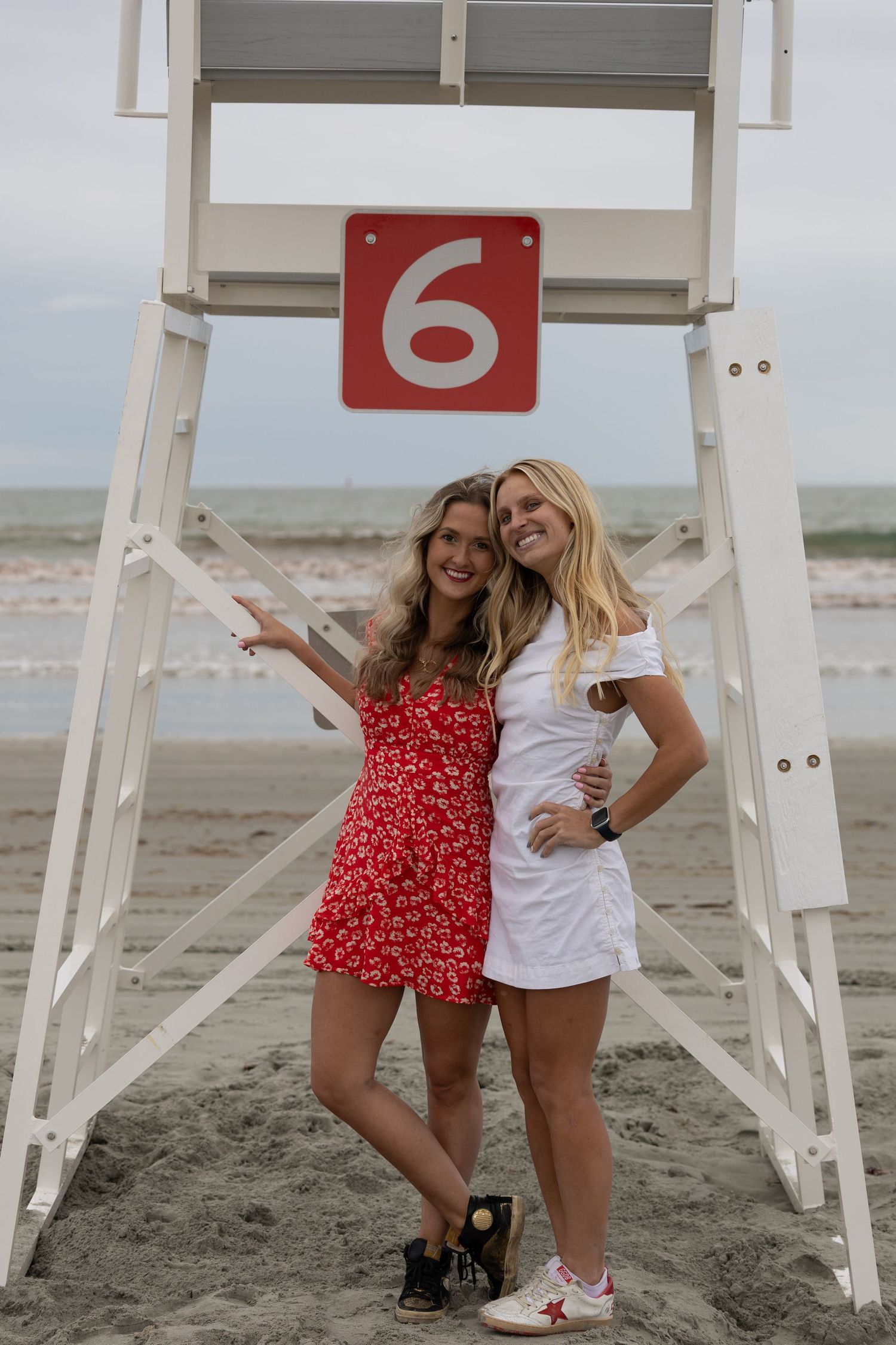 Two smiling women pose by lifeguard stand number 6 on a beach, one in red floral romper, one in white dress.