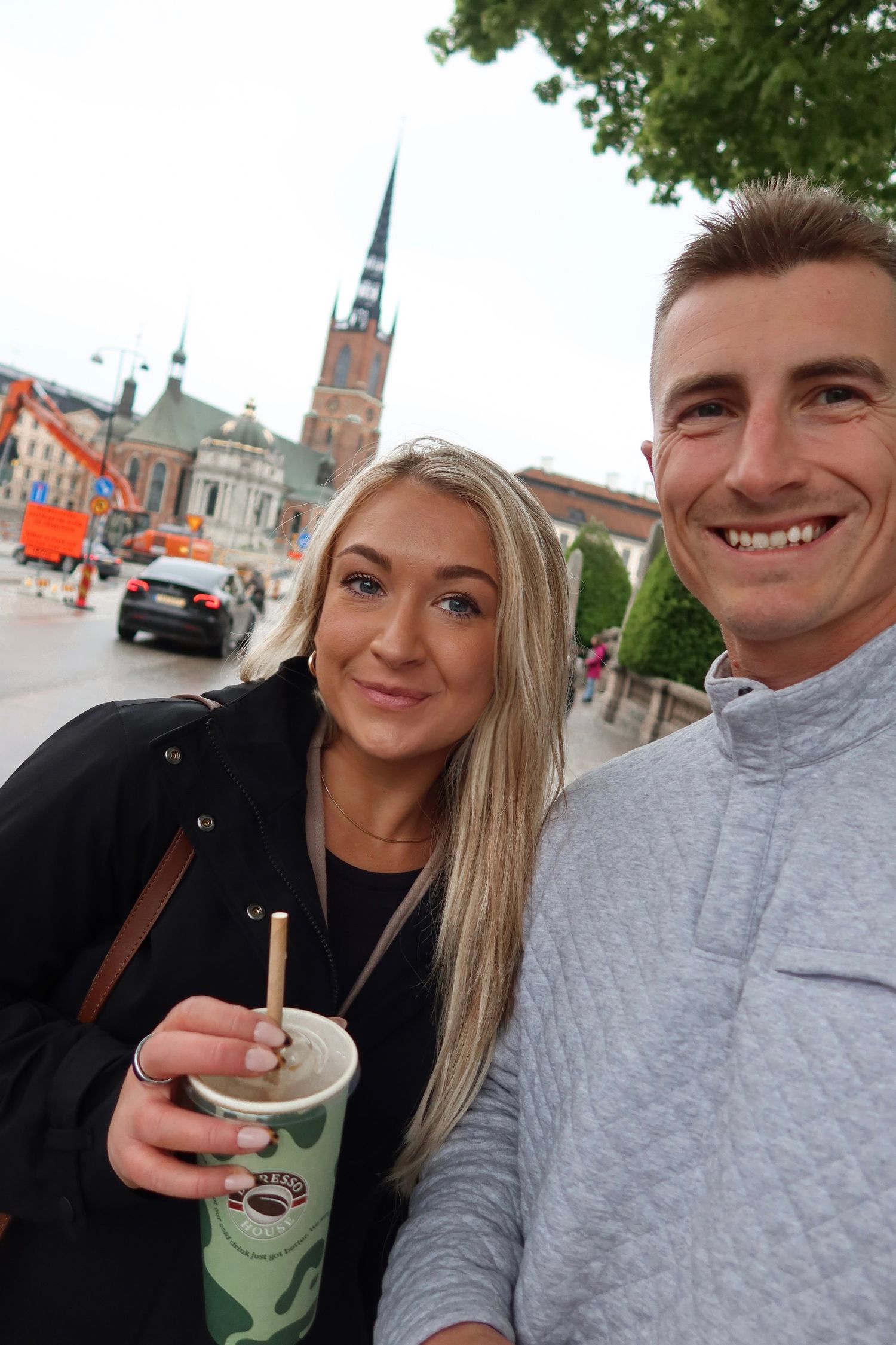 Two people smile while holding a drink in front of a historic church spire on a city street.