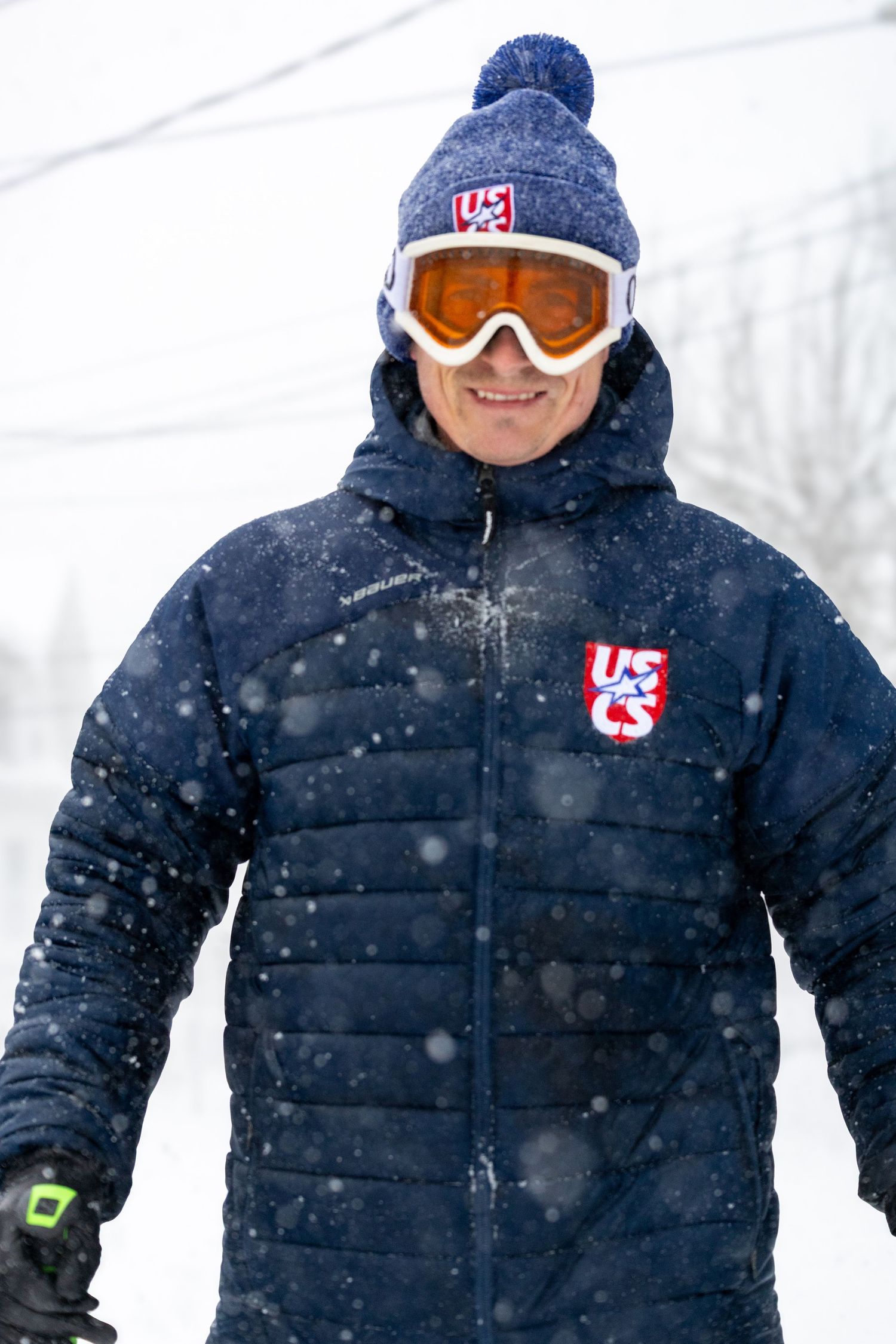 Skier wearing USA team winter gear including blue puffer jacket and knit hat stands in falling snow.