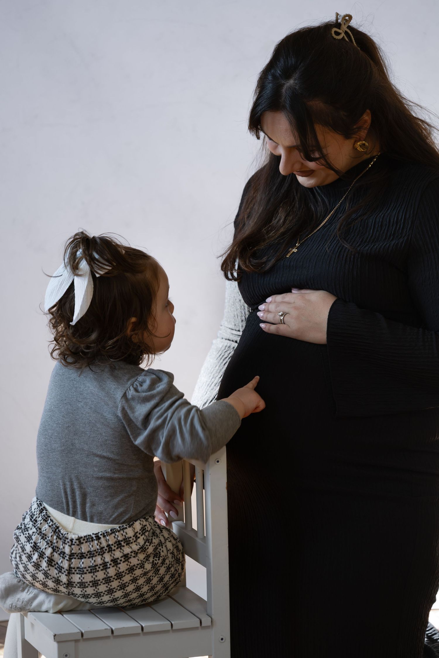 Pregnant woman in black dress smiling at toddler girl on white chair touching her baby bump.