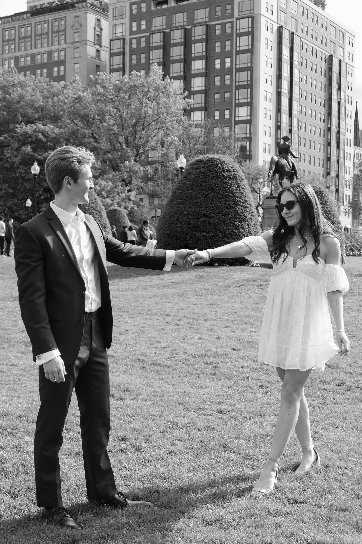 A couple in formal wear holds hands while dancing on grass with city buildings behind them.