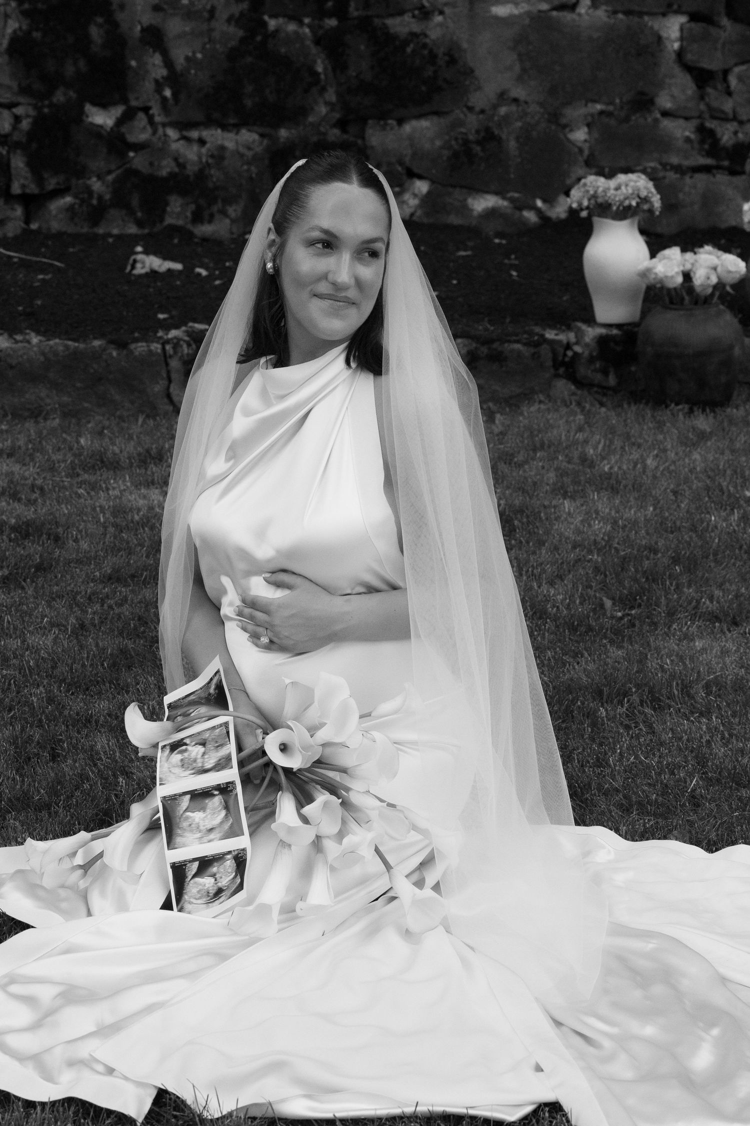 Pregnant bride in white silk gown and veil sitting on grass holding ultrasound photos and flowers.