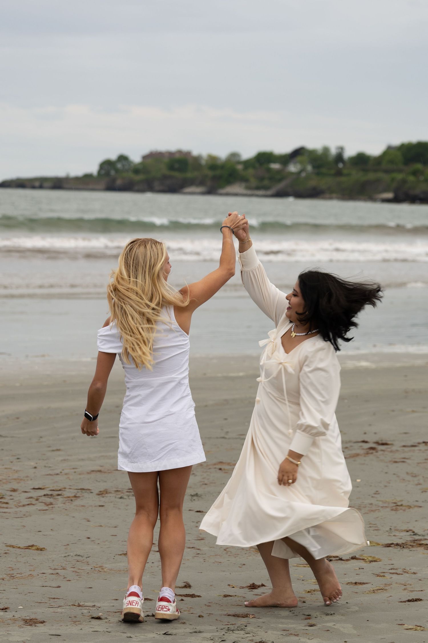 Two women in white outfits holding hands and spinning together on a scenic beach shoreline.