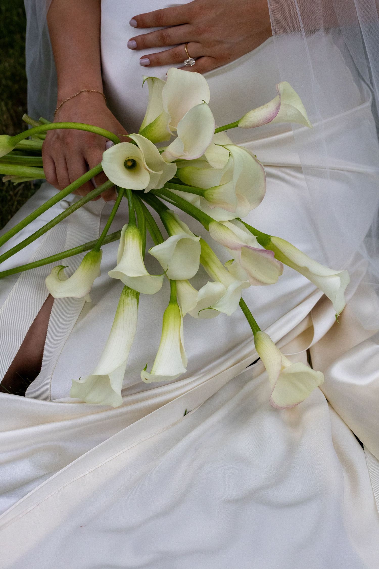 Bride holding elegant white calla lily bouquet against her flowing satin wedding dress, diamond ring visible.
