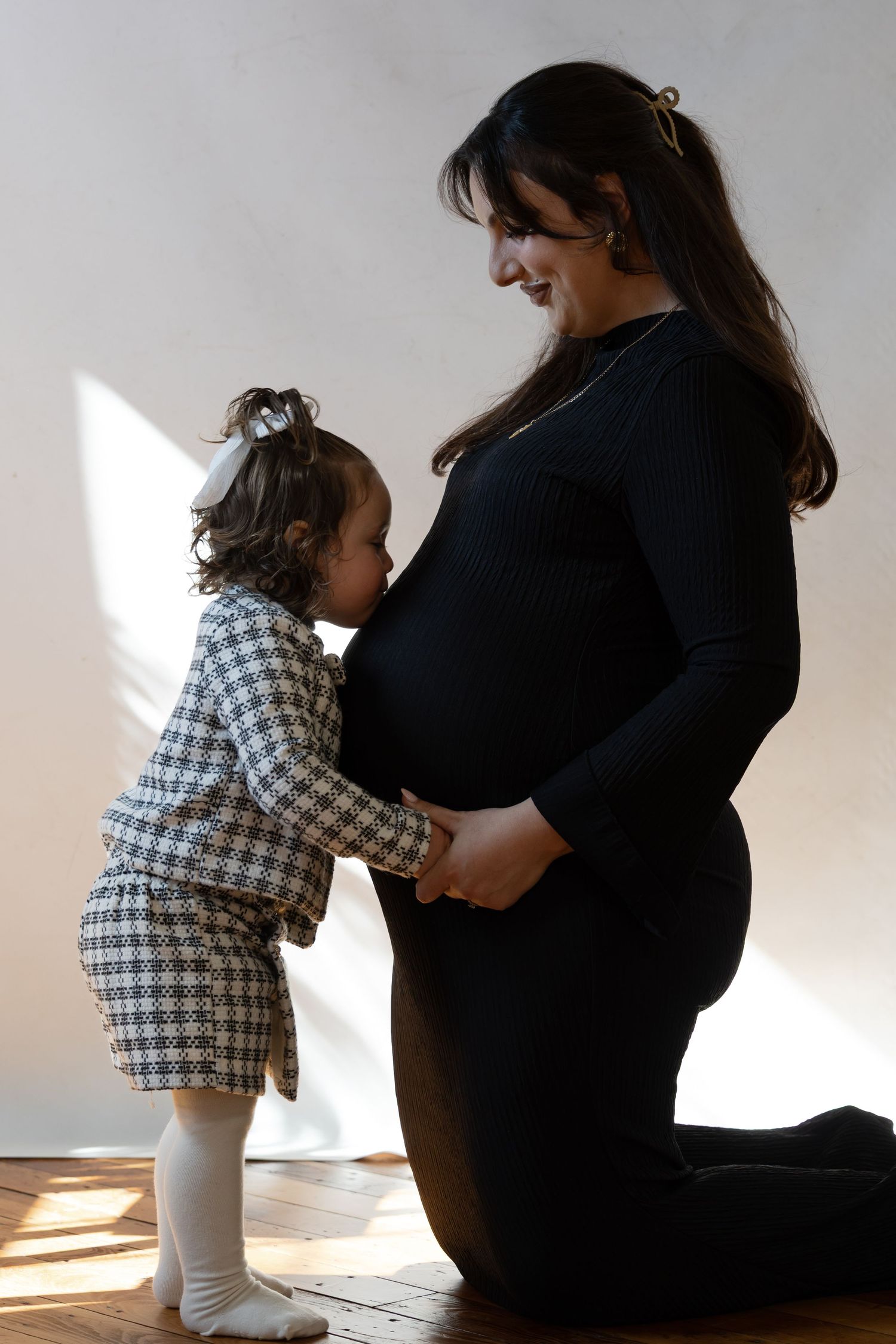 Pregnant woman in black outfit kneeling as toddler girl in plaid dress kisses her baby bump indoors.