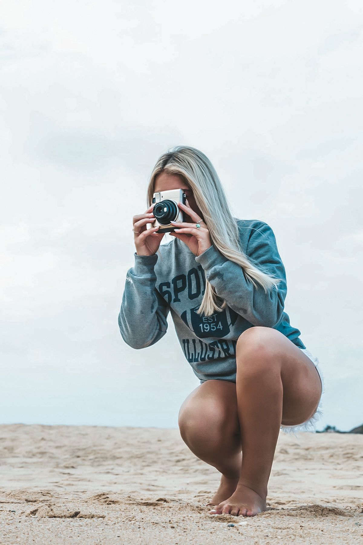 A photographer in a blue sweatshirt crouches on the beach taking photos with a professional camera.