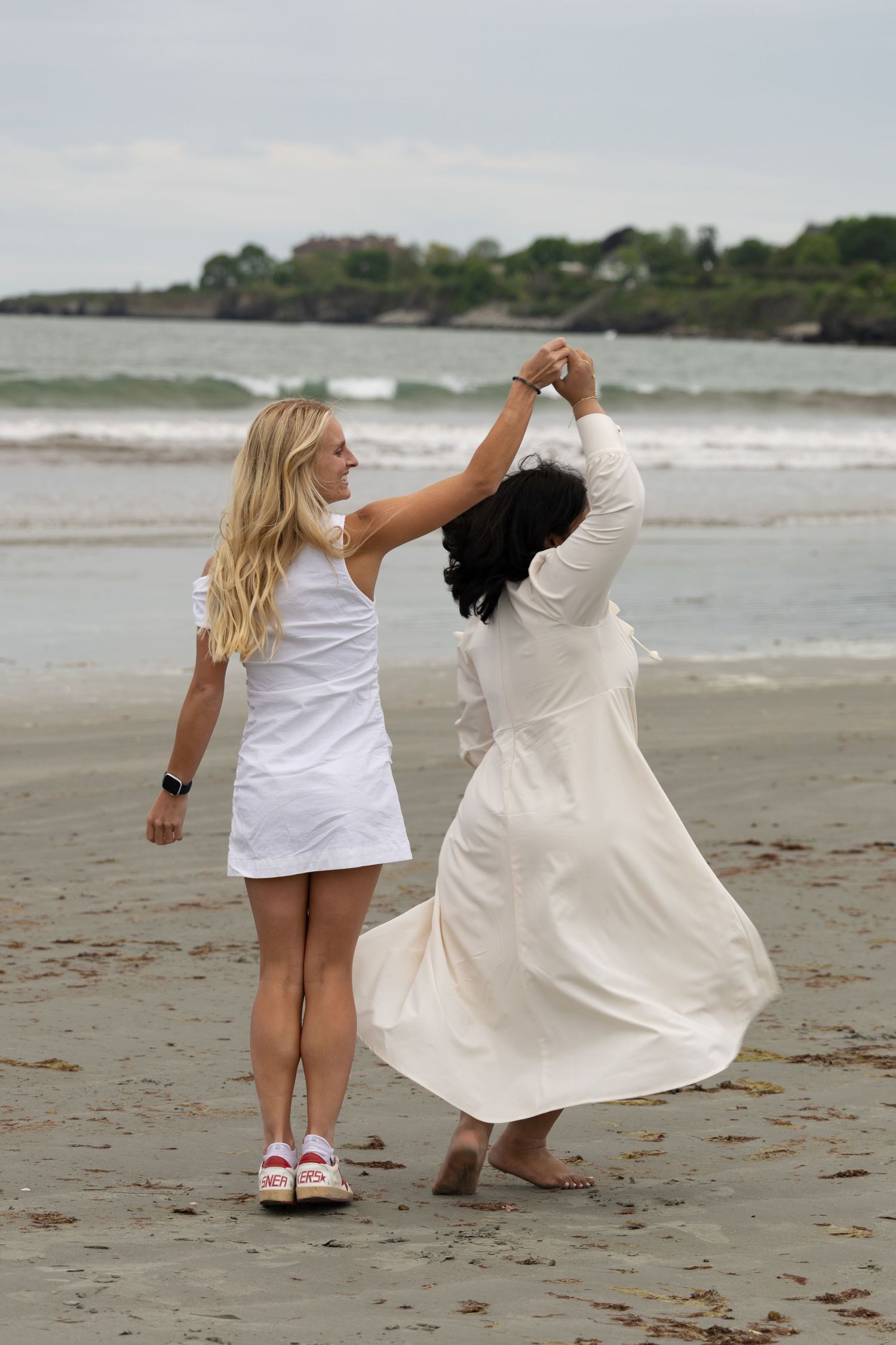 Two women in white dresses twirling together on a beach, viewed from behind with ocean scenery.