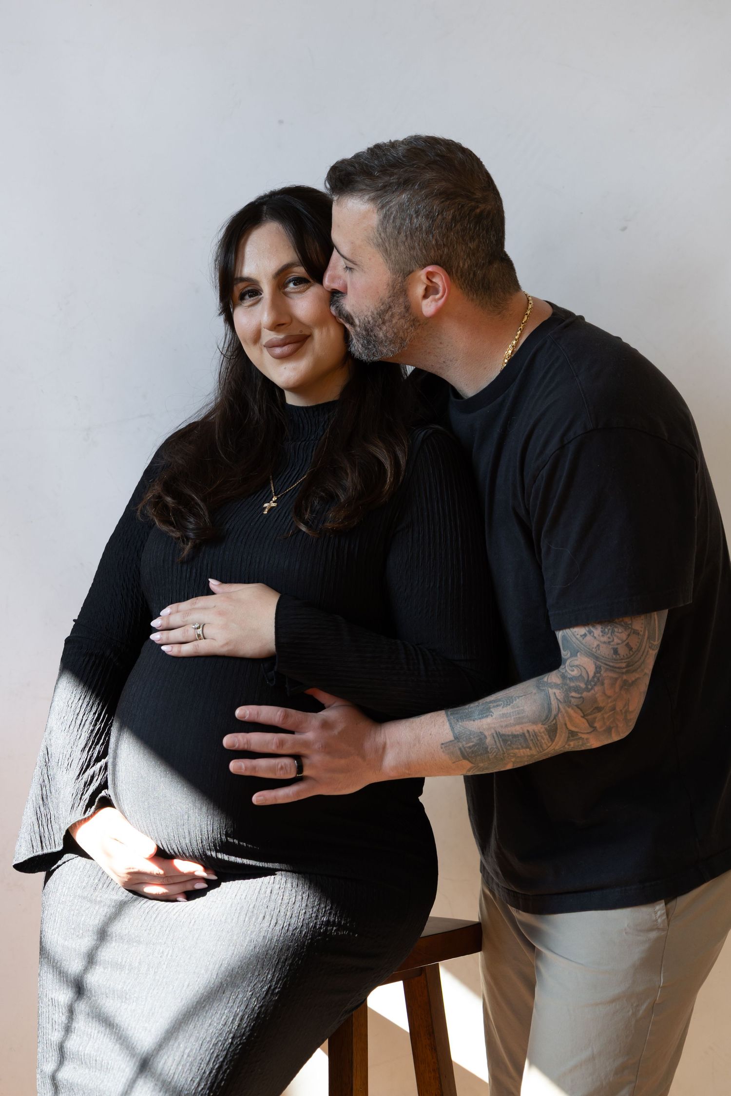 A man kisses a smiling pregnant woman on the cheek as they both cradle her baby bump in a studio.