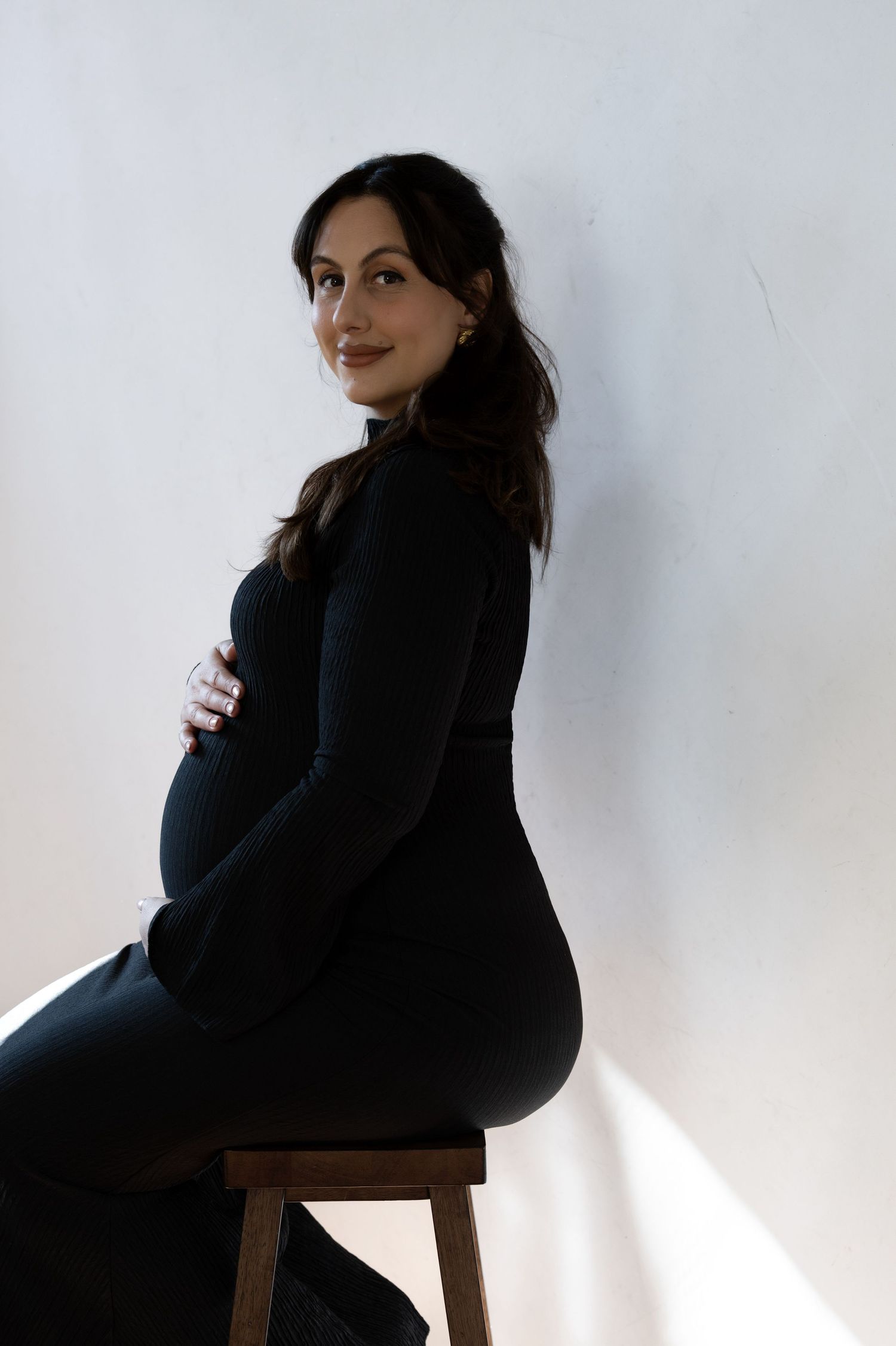 Pregnant woman in black outfit sitting on wooden stool, cradling baby bump against white studio background.