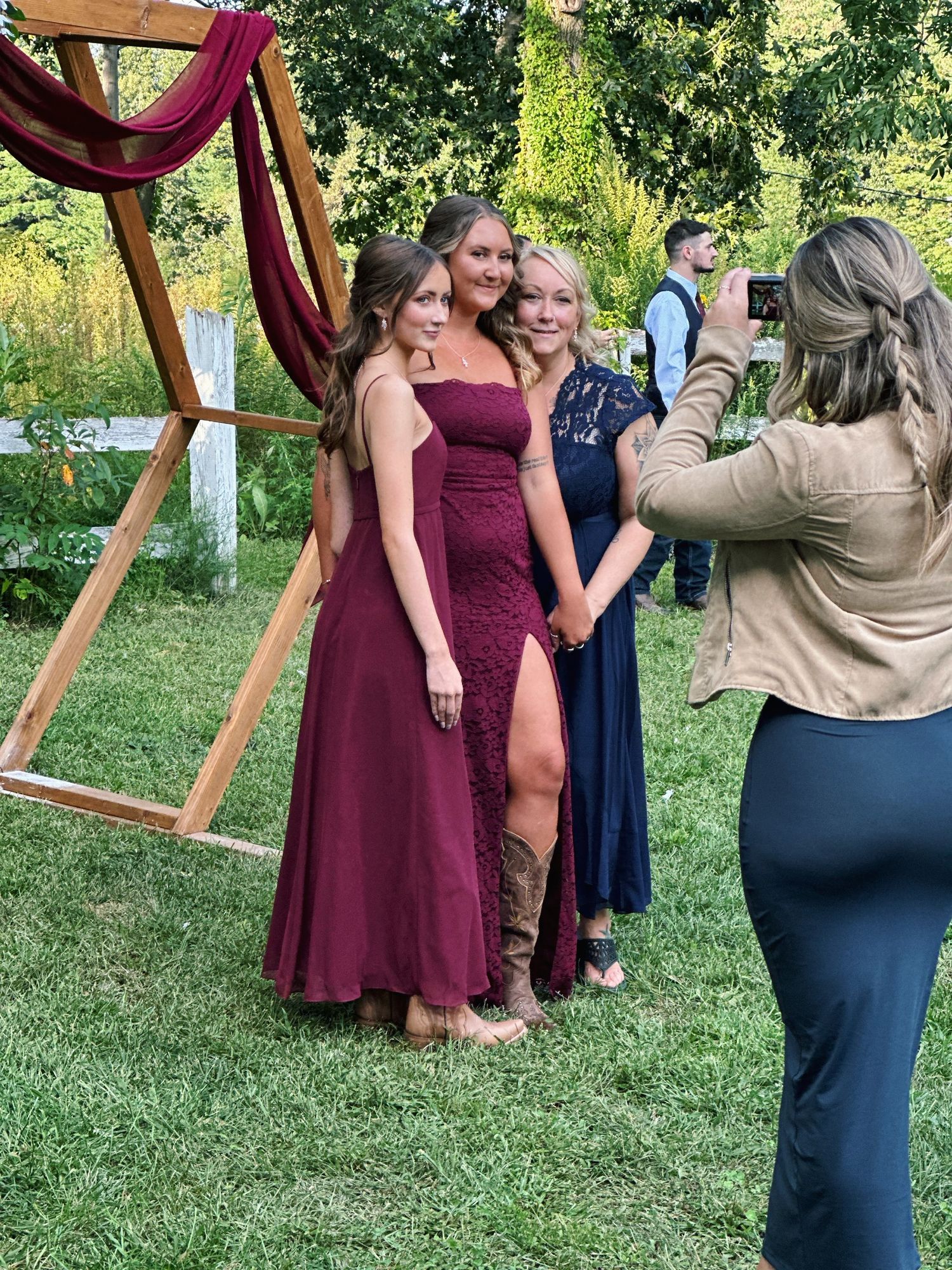 A group in burgundy and navy dresses poses playfully with a tan-suited figure in an outdoor garden setting.