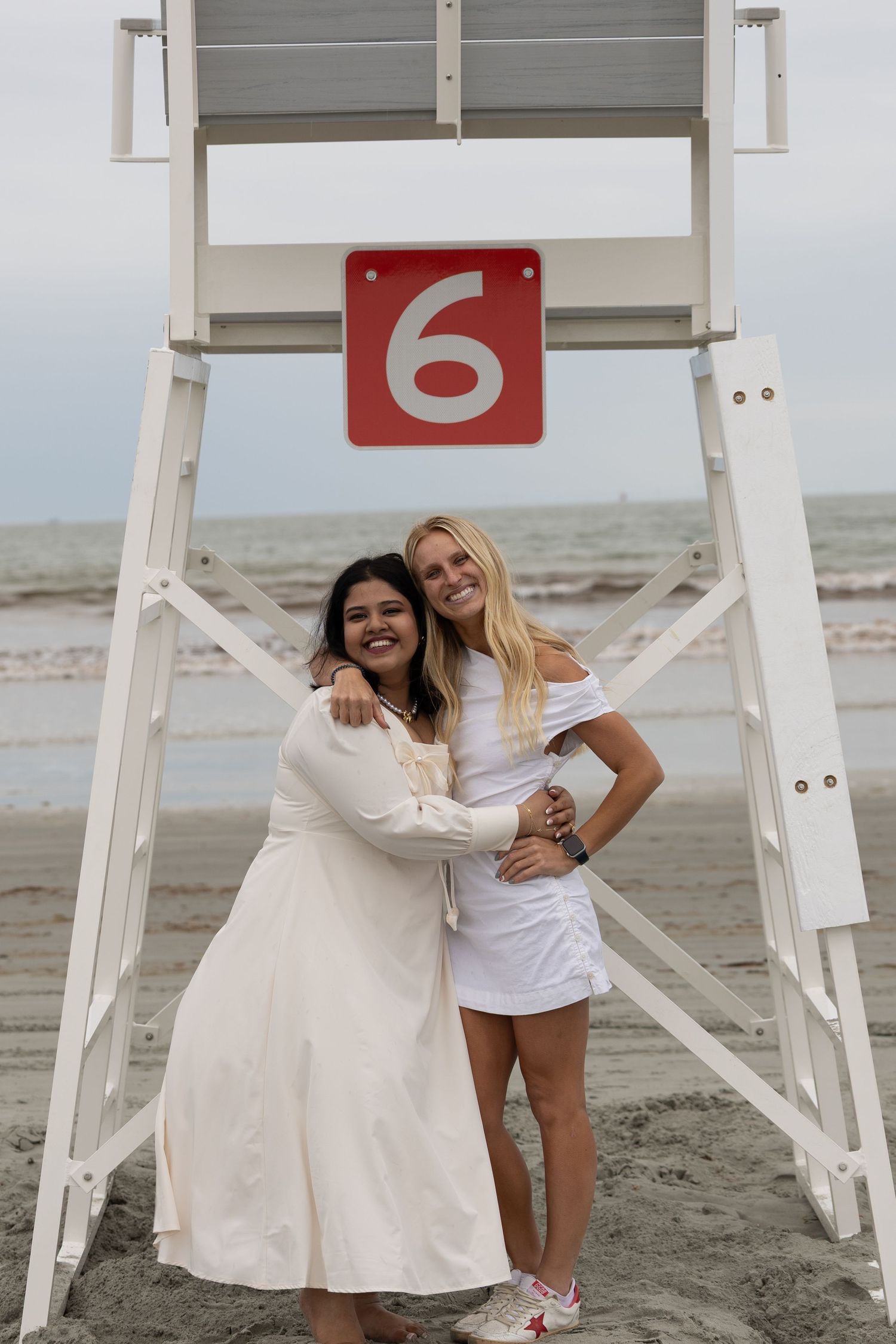 Two smiling women in white outfits embrace in front of lifeguard stand number 6 on a cloudy beach.