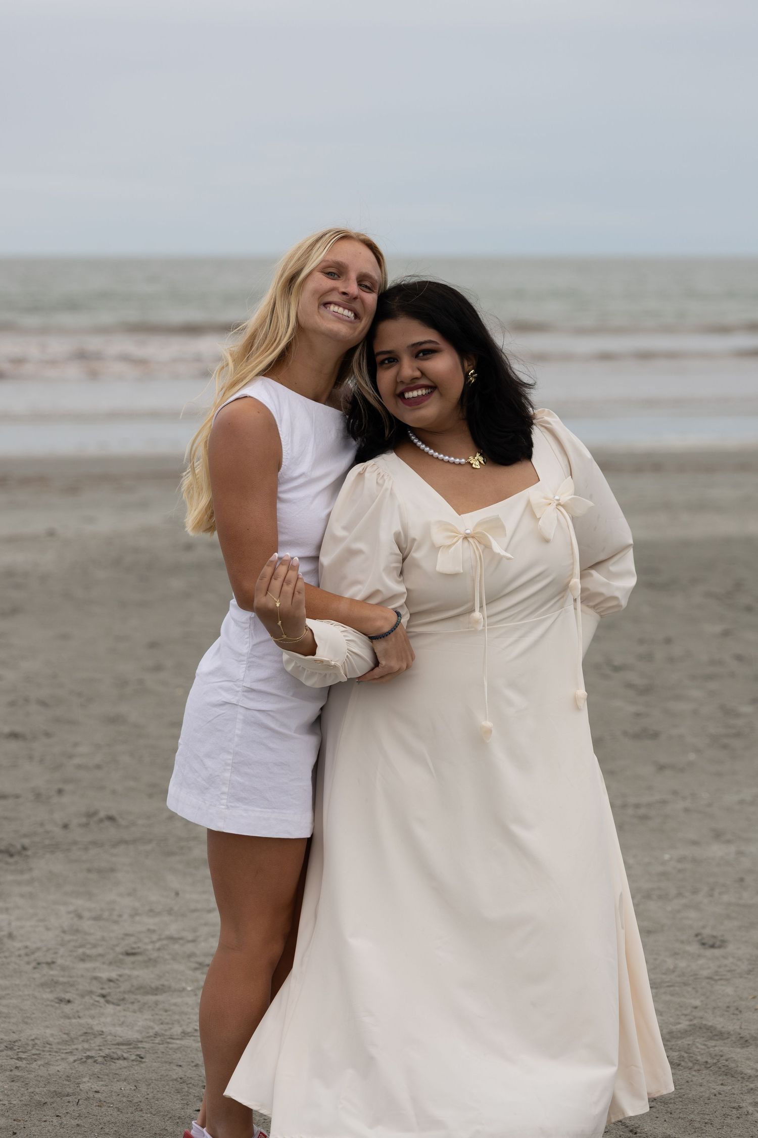 Two women in white dresses smiling and embracing on a beach with ocean in the background.